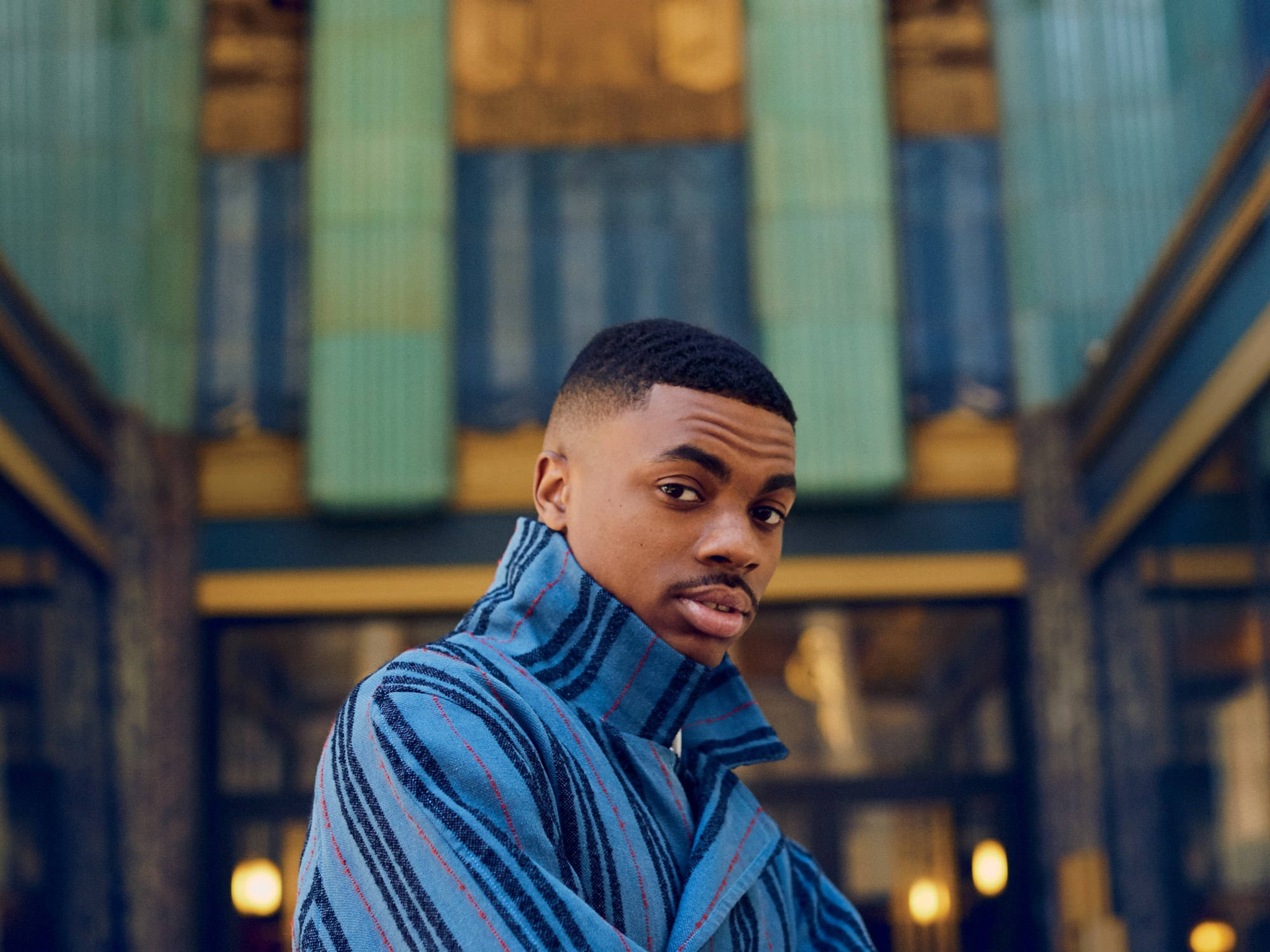 酷炫Vince Staples 壁紙 — 一名身穿藍色條紋外套的年輕男子在色彩繽紛的建築物外牆前直視鏡頭。