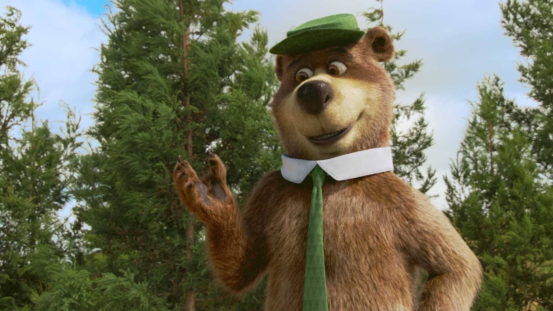 酷炫Yogi Bear場景 壁紙 — 一隻戴著綠色帽子和領帶的開心動畫熊在明亮的天空下，在高大的松樹間愉快地揮手。
