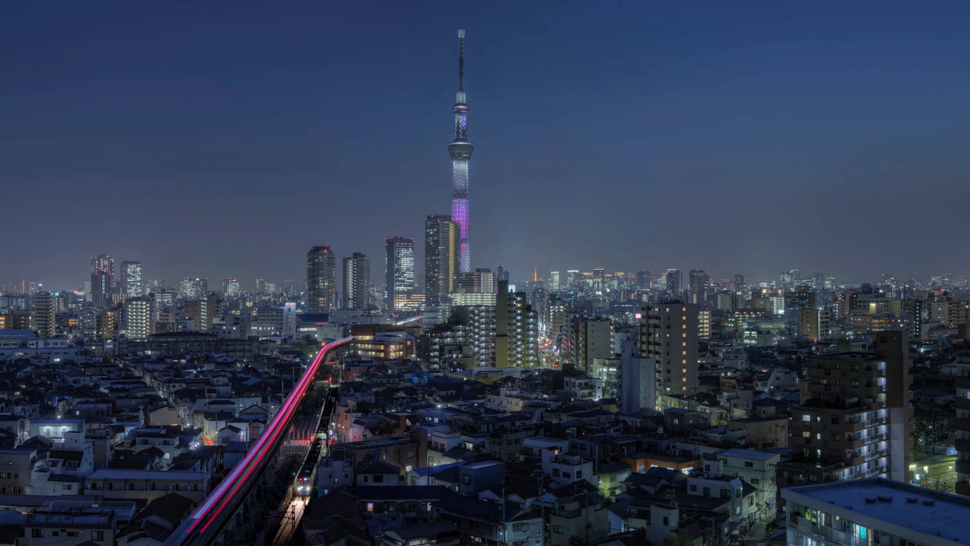 酷炫日本新宿 壁紙 — 夜晚的東京天際線，東京晴空塔發出紫色和粉紅色的光芒，以及一列經過的火車留下的光跡。