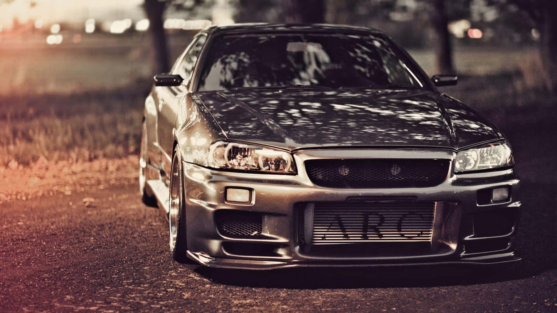銀色日產Skyline R34在黃金時刻停泊在碎石路上，背景有樹木和城市燈光。