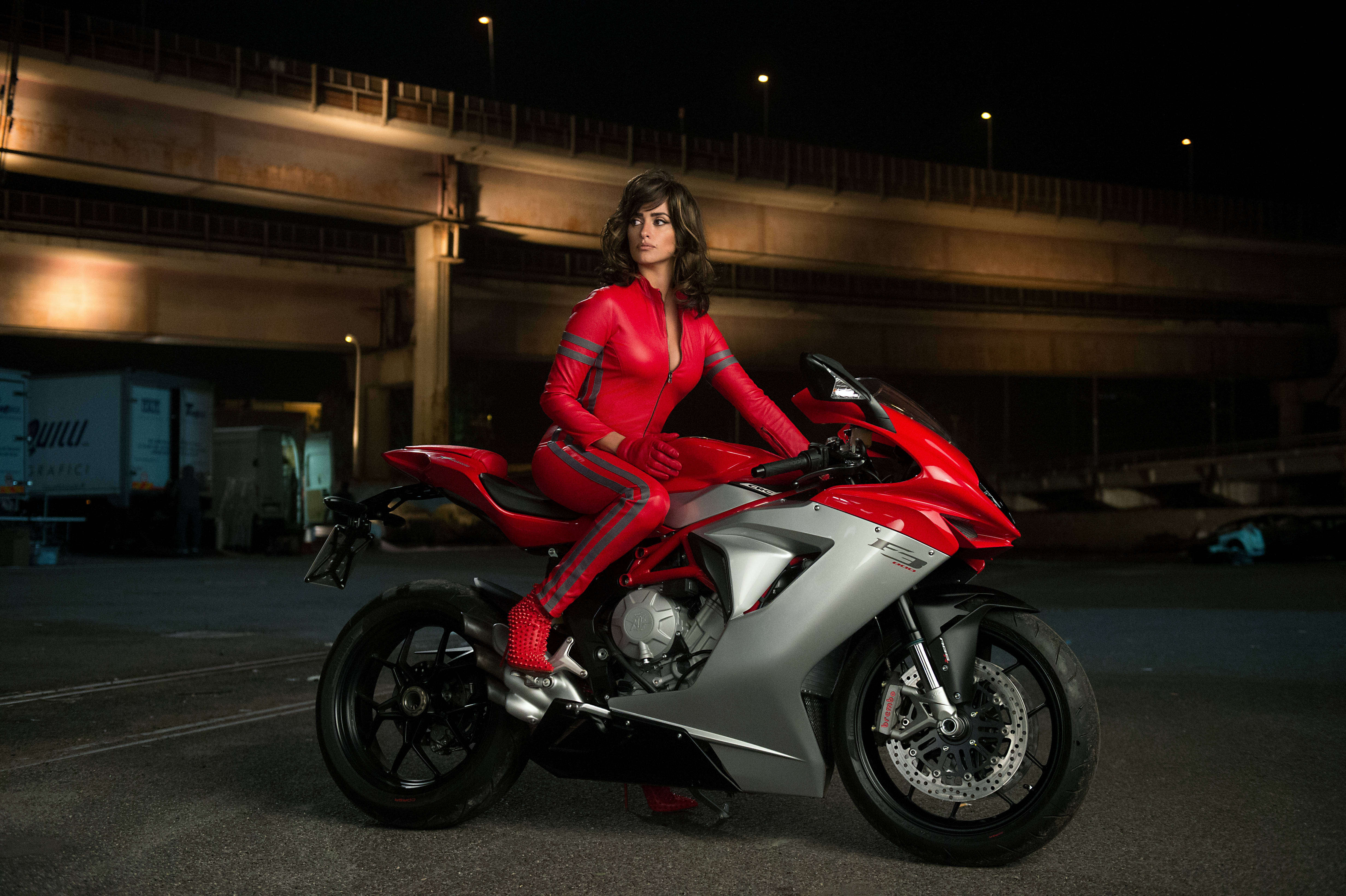 酷炫機車騎士佩內洛普·克魯茲 4K 壁紙 — 一名身穿紅色賽車服的女士在夜間坐在一輛紅銀色運動電單車上，位於有燈光的天橋下。
