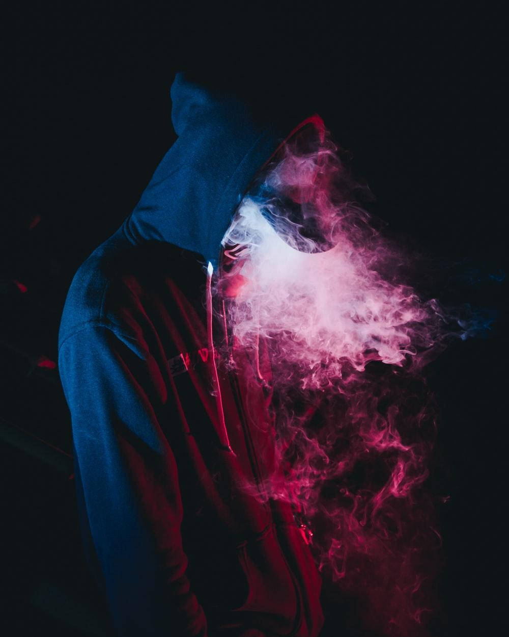 酷炫煙霧氛圍 壁紙 — 一個戴著連帽衫的人物在黑暗背景下呼出鮮豔的粉紅色和白色蒸氣，背景被霓虹藍和紅燈照亮。