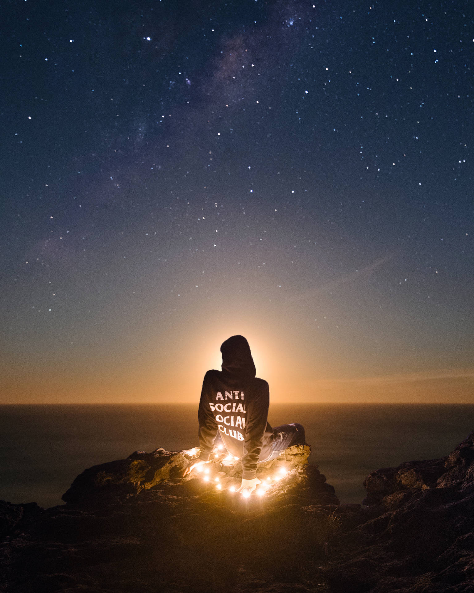 穿連帽衫的人夜晚坐在懸崖上，有串燈，觀看海洋上空的星星