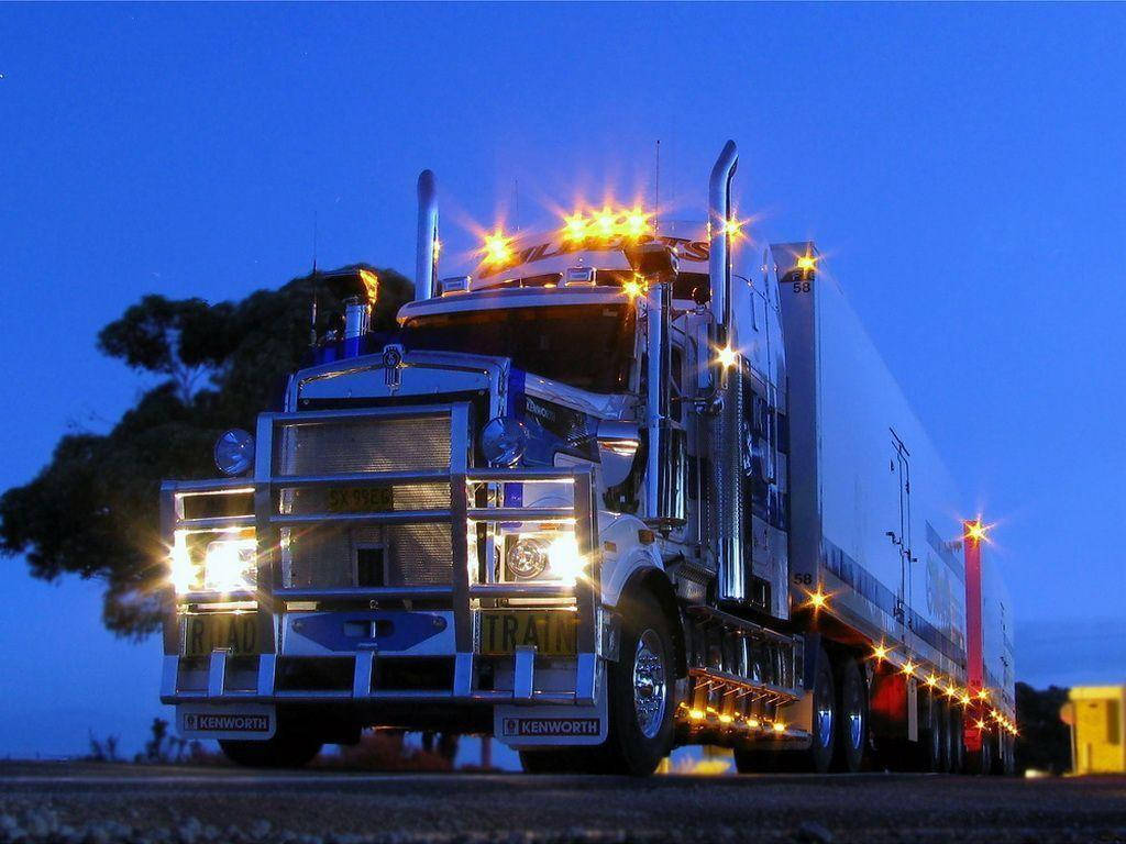 酷炫肯沃斯卡車 壁紙 — 一輛閃亮的白色Kenworth半掛卡車，明亮的車頭燈和琥珀色警告燈照亮了黑暗樹木旁的暮色道路。