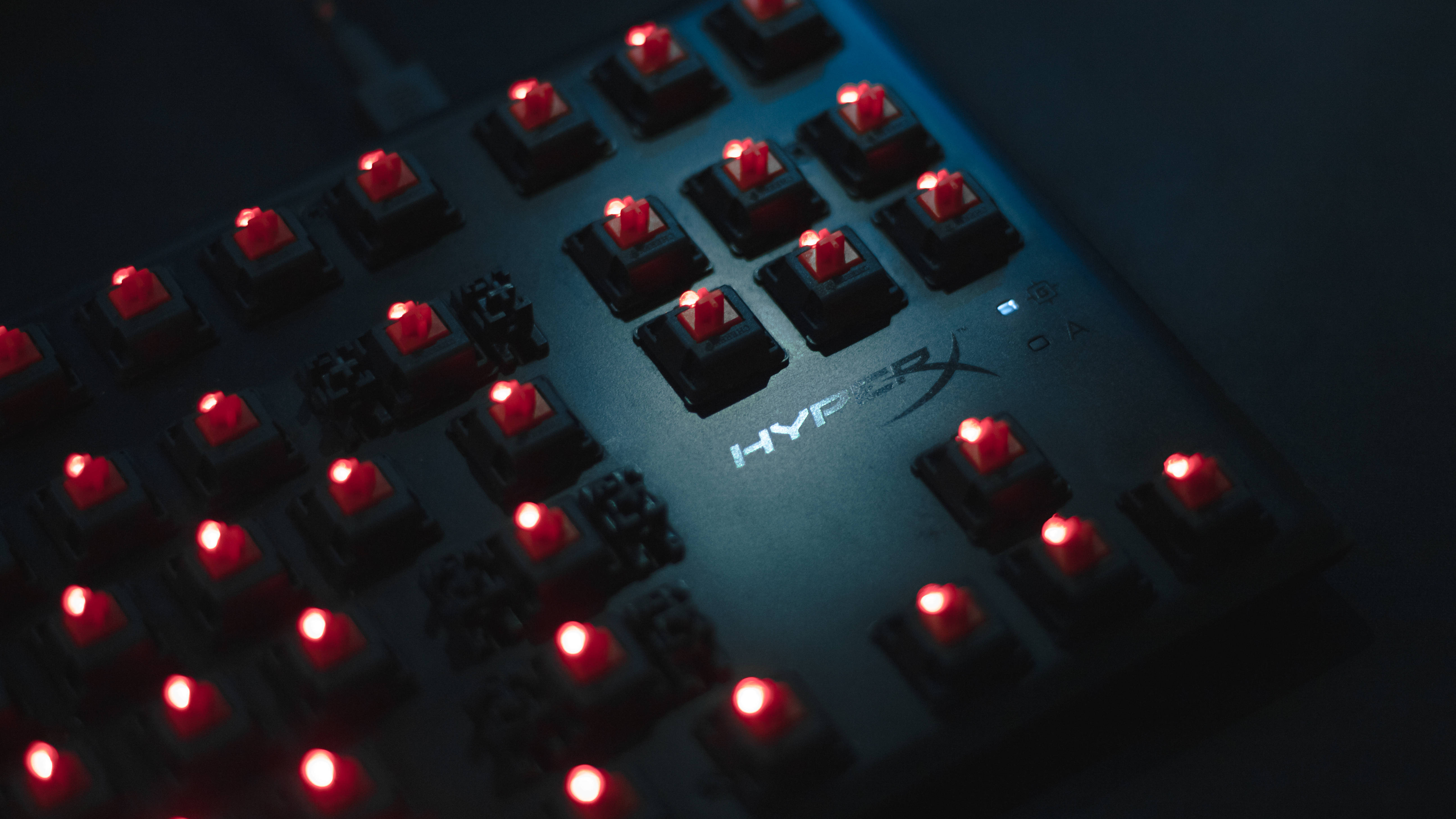 酷炫遊戲HyperX鍵盤 壁紙 — 近距離拍攝的遊戲鍵盤，帶有紅色背光和HyperX品牌標誌，處於昏暗燈光下。