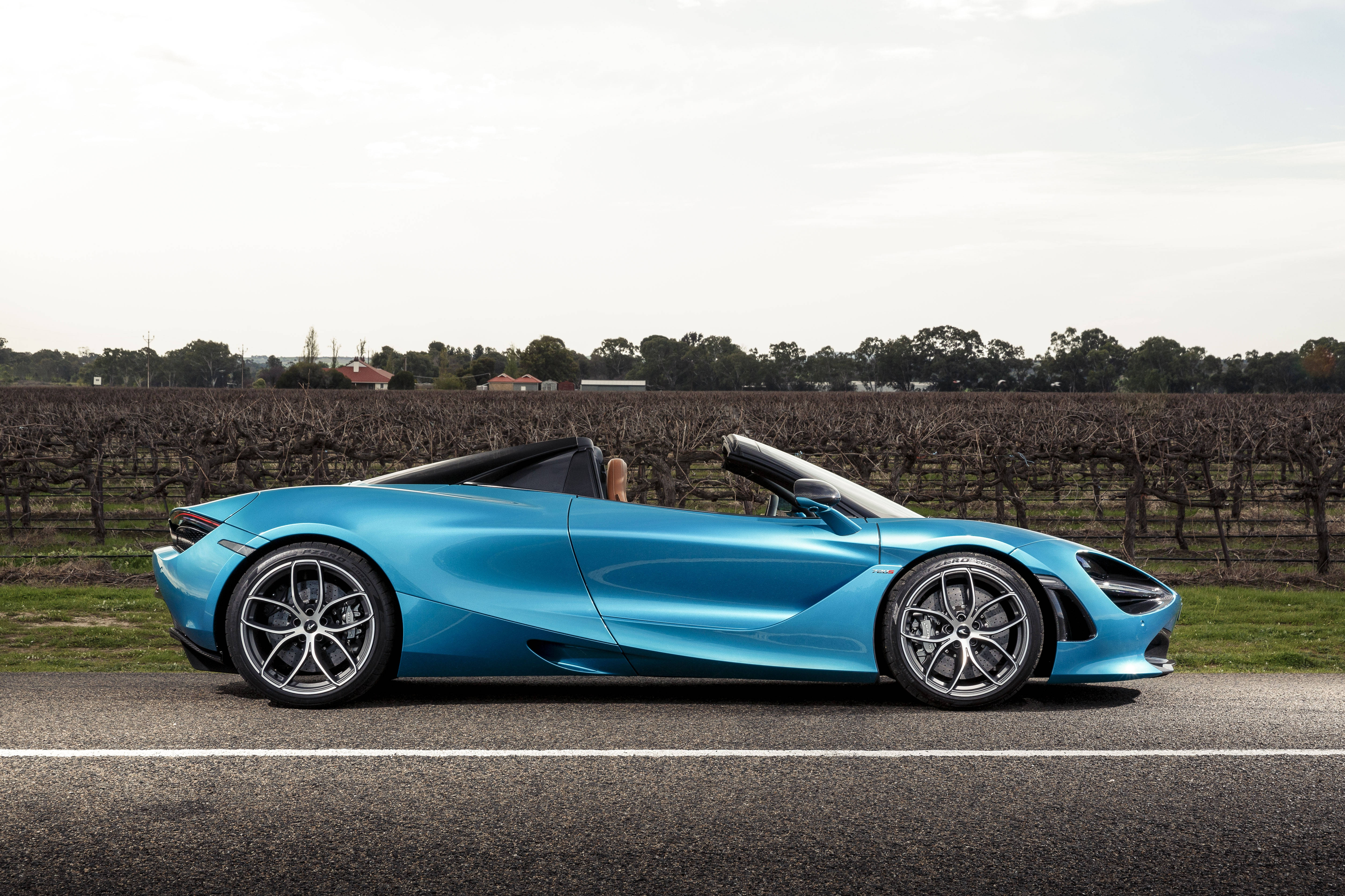 酷藍色敞篷麥拉倫720S 4K 壁紙 — 一架時尚的藍色開篷跑車在陰天下行駛在葡萄園旁邊的鄉村道路上。