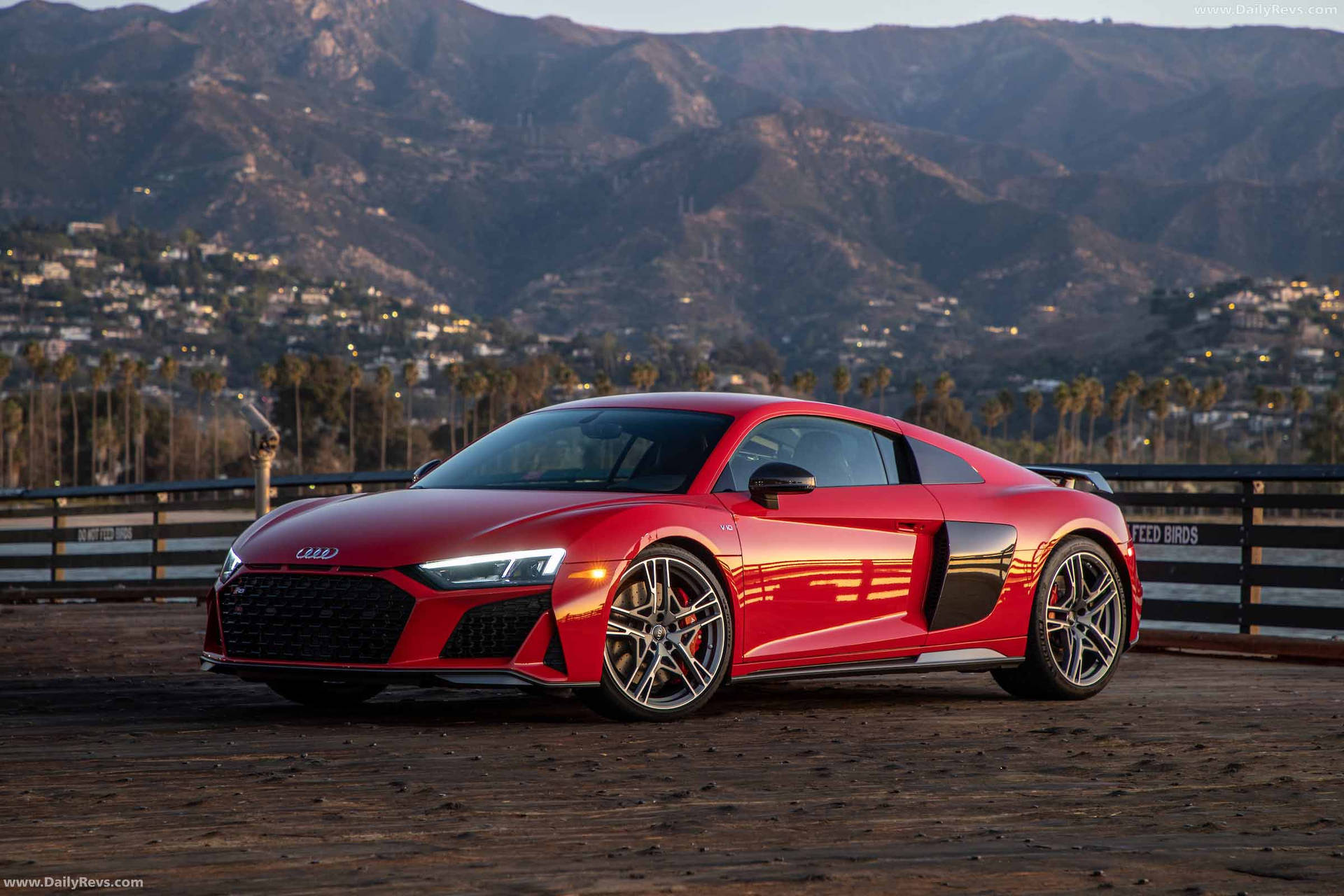 日落時分，紅色Audi R8跑車停在木製碼頭，背景有山脈和沿海小鎮。