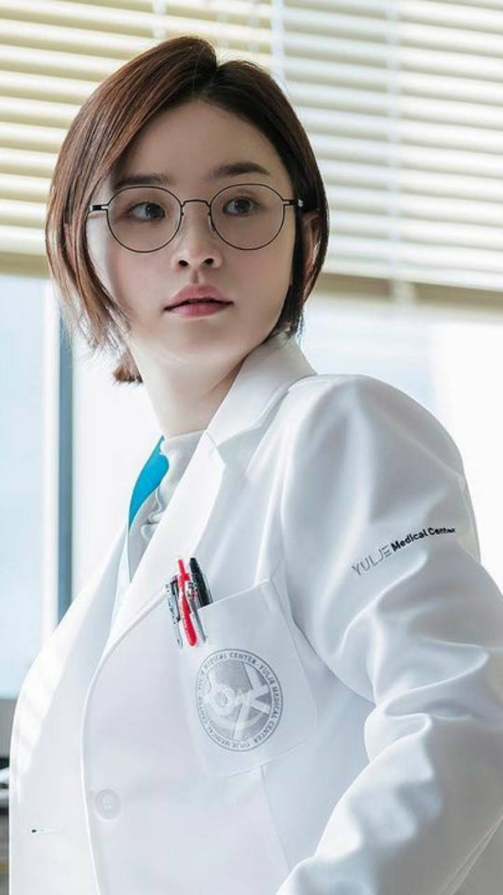 《醫院播放清單》成惠白袍 壁紙 — 一位年輕女醫生穿著潔白的實驗室外套，站在有百葉窗的明亮辦公室裡，表情嚴肅地直視鏡頭。