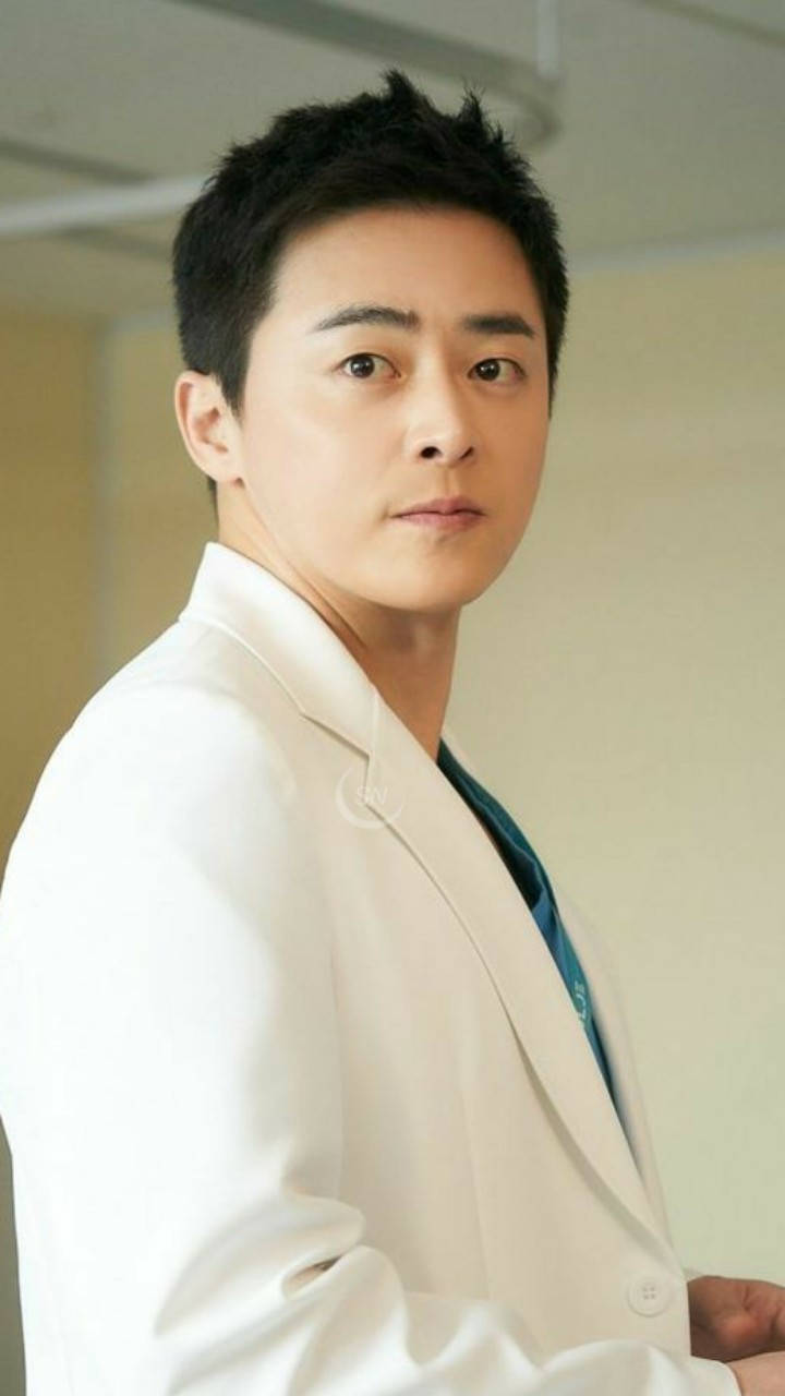 《醫院播放清單》李益俊肖像 壁紙 — 一位年輕男子穿著俐落的白色西裝外套和青綠色襯衫，在柔和光線的室內環境中，表情嚴肅地回頭看。