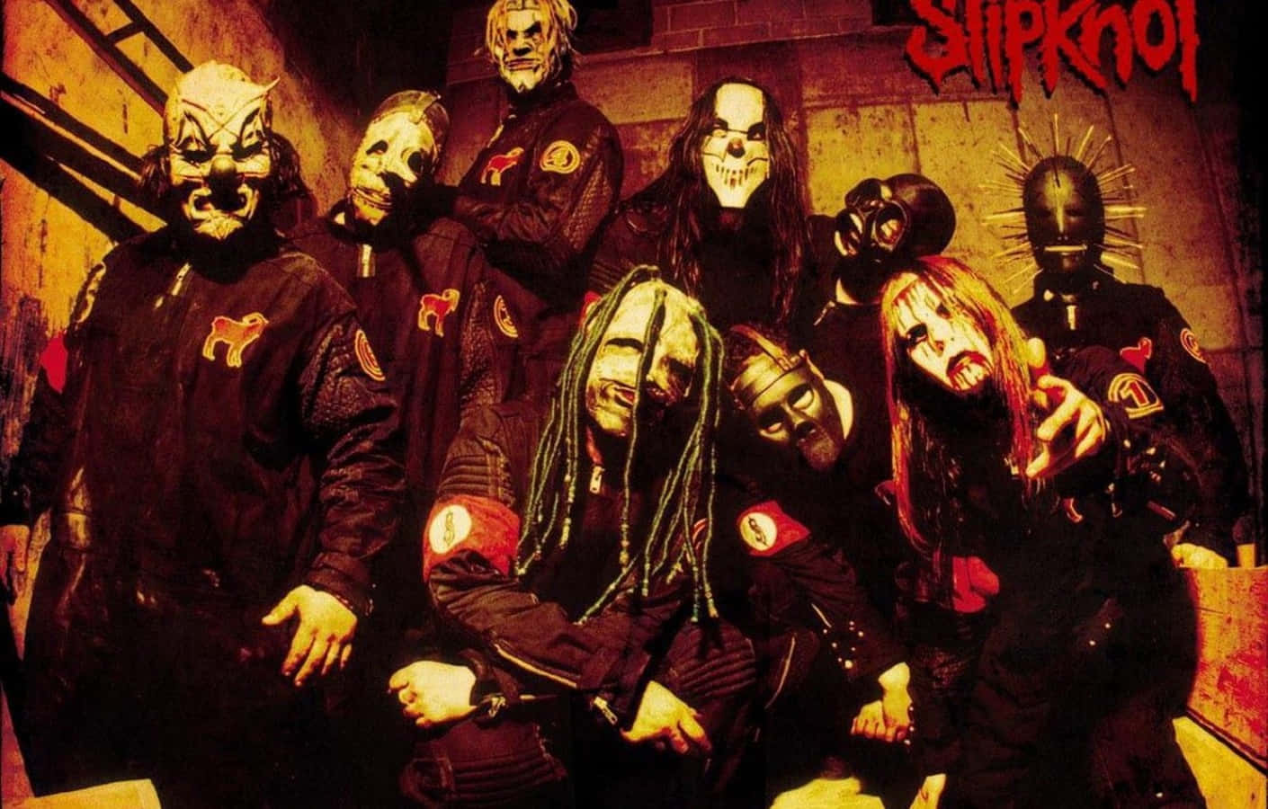 釋放你的黑暗面，使用Slipknot桌面 壁紙 — 七位戴面具的音樂家身穿黑色外套，在溫暖的橙色燈光下，在粗糙的磚牆前擺出強烈的姿勢。