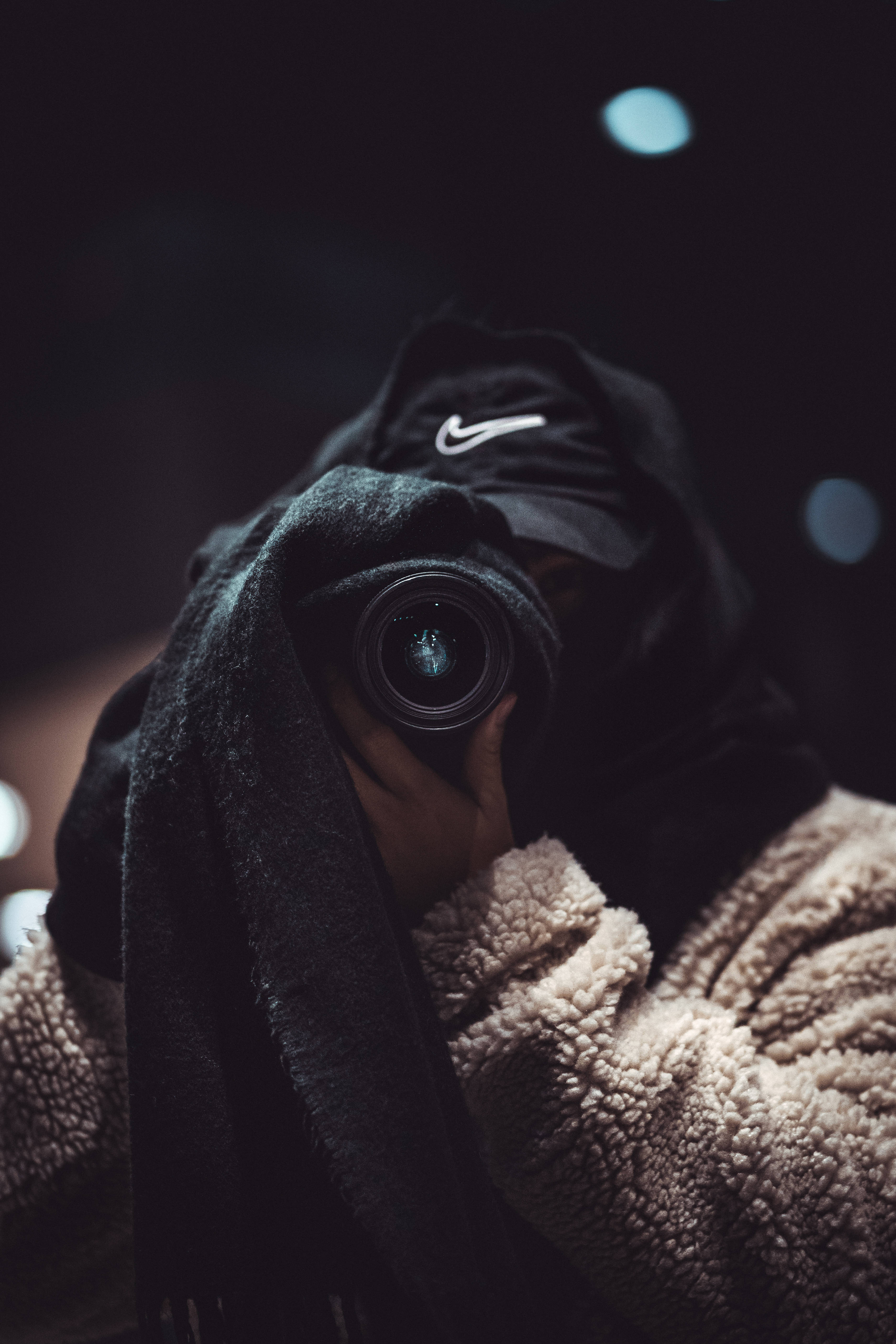 釋放速度 - 4K Nike 鞋藝術 壁紙 — 一個穿著黑色連帽衫和毛絨米色外套的人，在帶有柔和藍色散景燈光的黑暗背景下將相機舉到臉前。