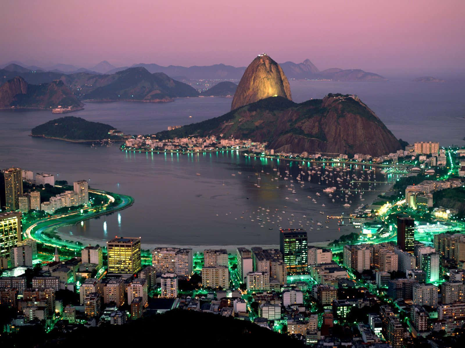 Brazil 桌布 壁紙 — 里約熱內盧黃昏時分的航拍圖，糖麵包山在粉紅色天空下閃耀，城市燈光倒映在水面上。
