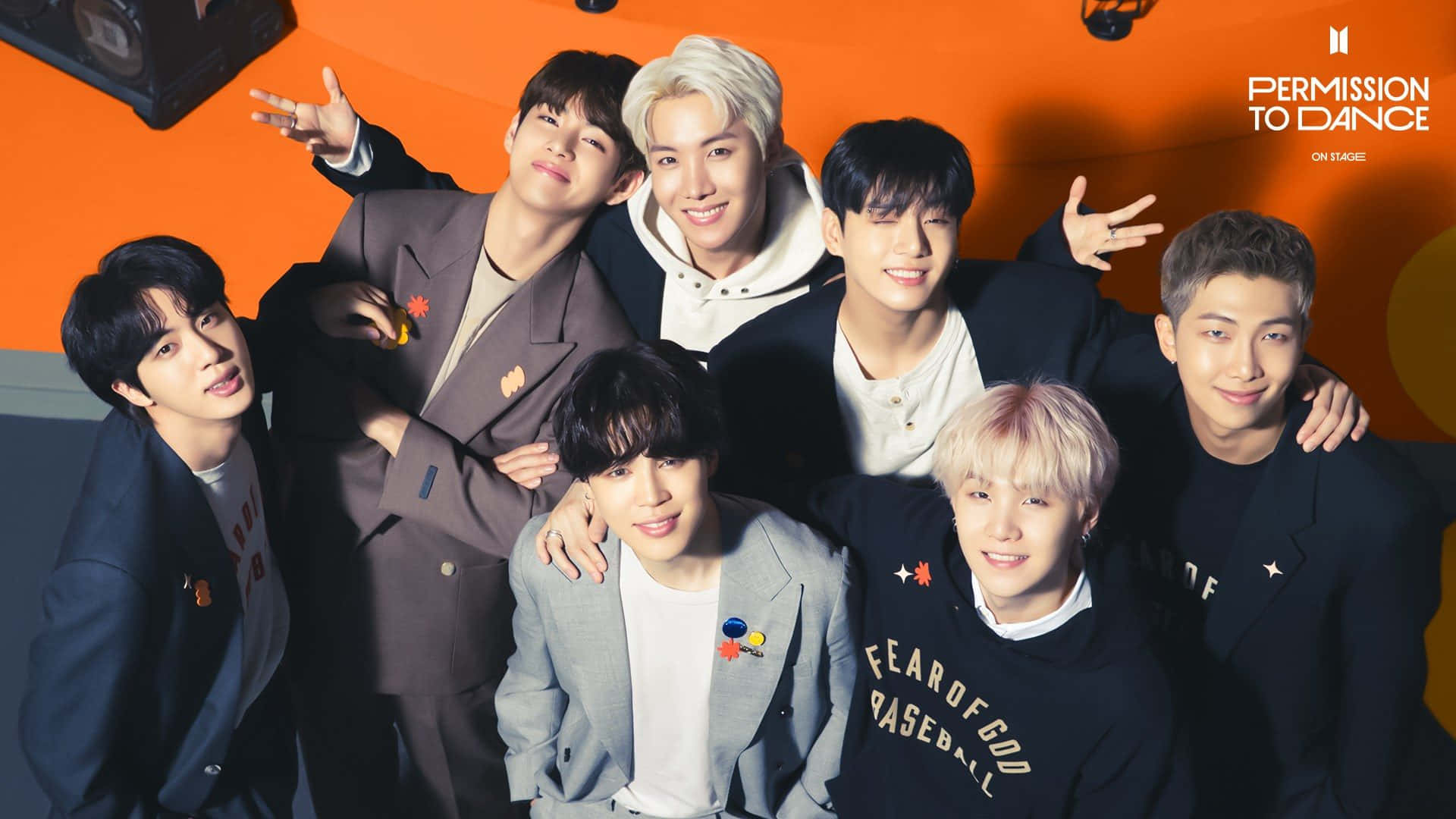 七位K-pop偶像穿著時尚服裝，在橙色背景的舞台上一起擺姿勢。