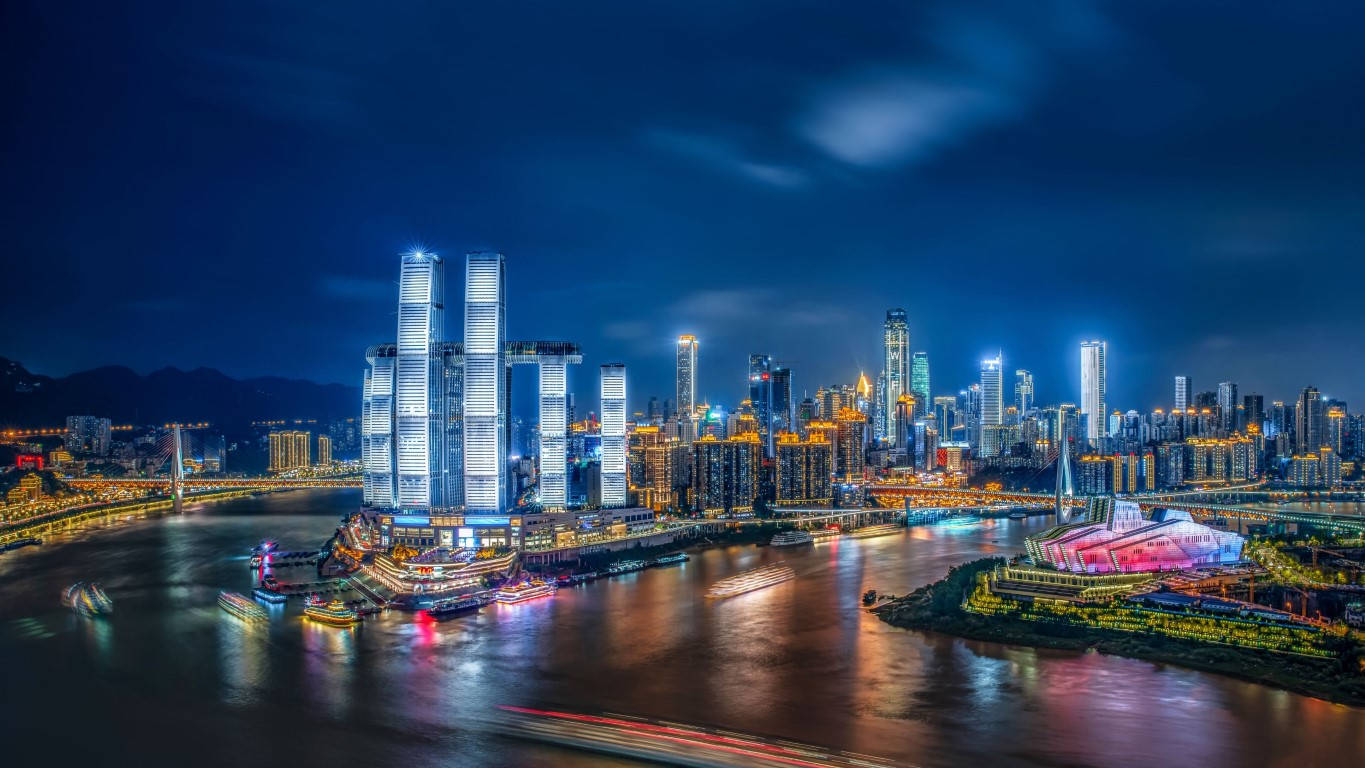 重慶中國摩天大樓城市風景 壁紙 — 令人目眩的夜景城市景觀，燈火通明的摩天大樓倒映在河面上，伴隨著船隻留下的光跡。