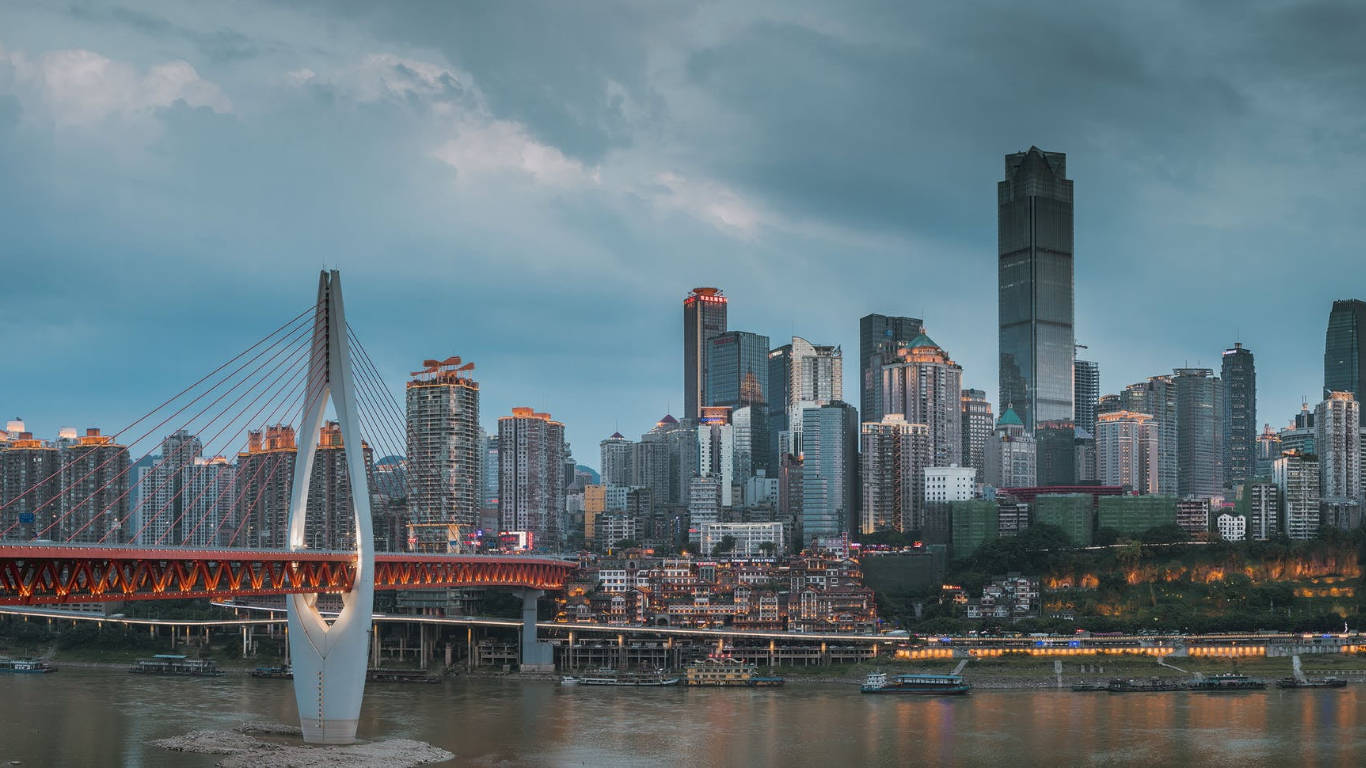 重慶中國都市天際線 壁紙 — 黃昏時分的現代城市景觀，一座發光的紅色橋樑橫跨河流，摩天大樓倒映在水中。