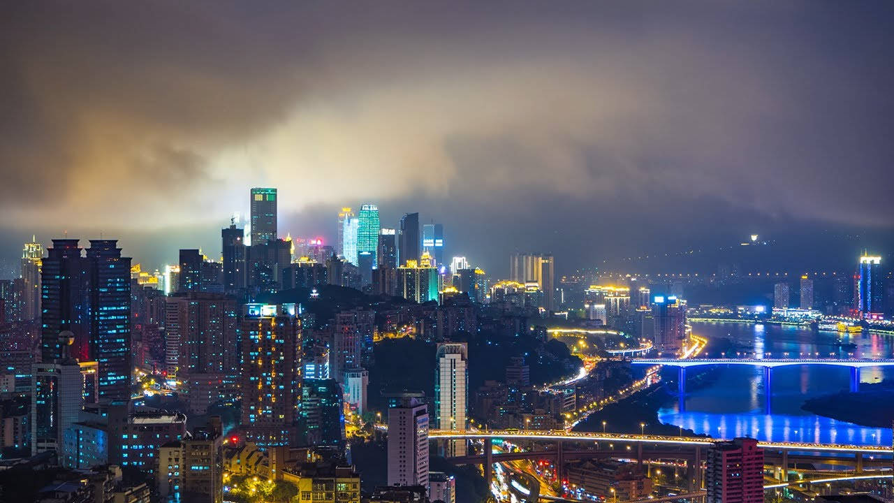 重慶城市夜景霓虹燈光 壁紙 — 一個充滿活力的夜景城市景觀，在戲劇性的多雲天空下，河流上有發光的摩天大樓和橋樑。