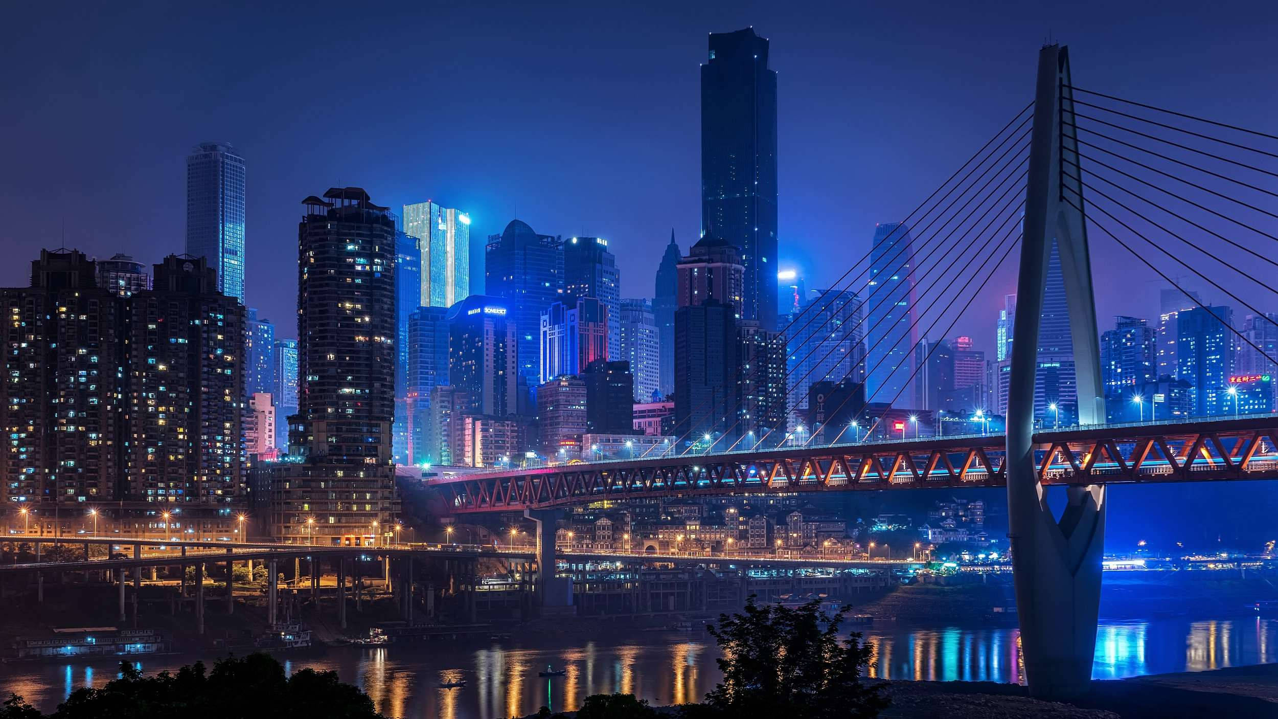 重慶夜間美學風景 壁紙 — 充滿活力的夜景城市景觀，一座發光的橋樑橫跨河流，反射著高聳摩天大樓的霓虹燈光。