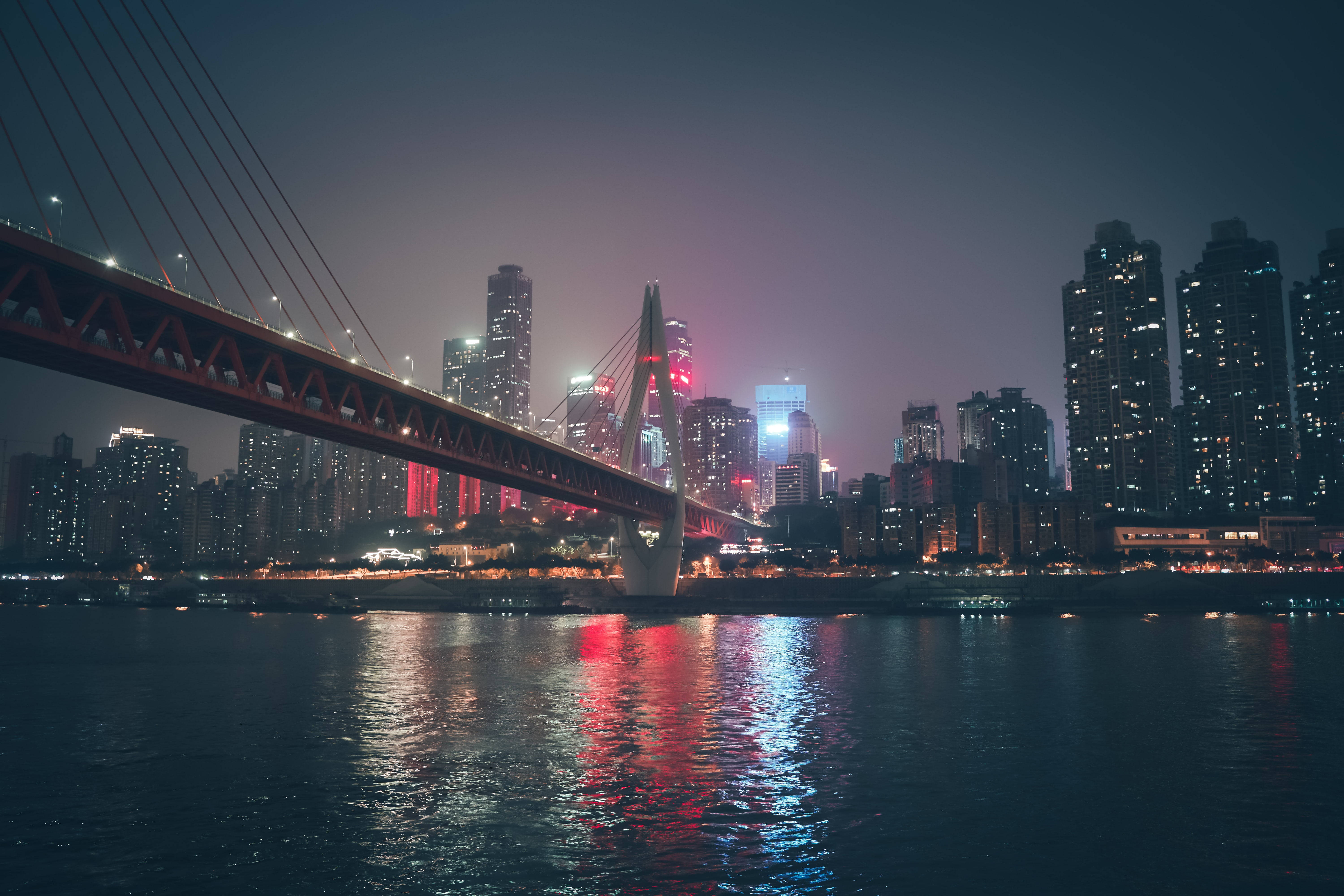 重慶橋樑夜景 壁紙 — 一座發光的紅色吊橋橫跨黑暗的河流，映照著霓虹城市燈光，背景是朦朧的夜空。