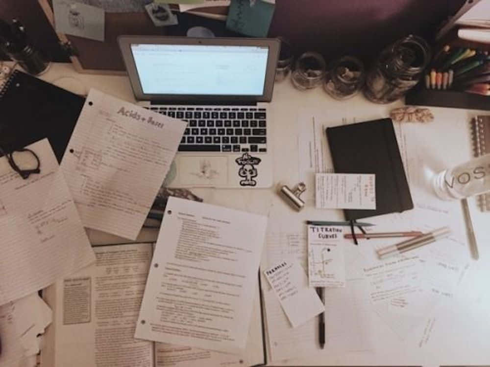 重拾學習的熱情 壁紙 — 在溫暖的環境光線下，俯瞰一張雜亂的書桌，上面散落著化學筆記、手提電腦和文具。