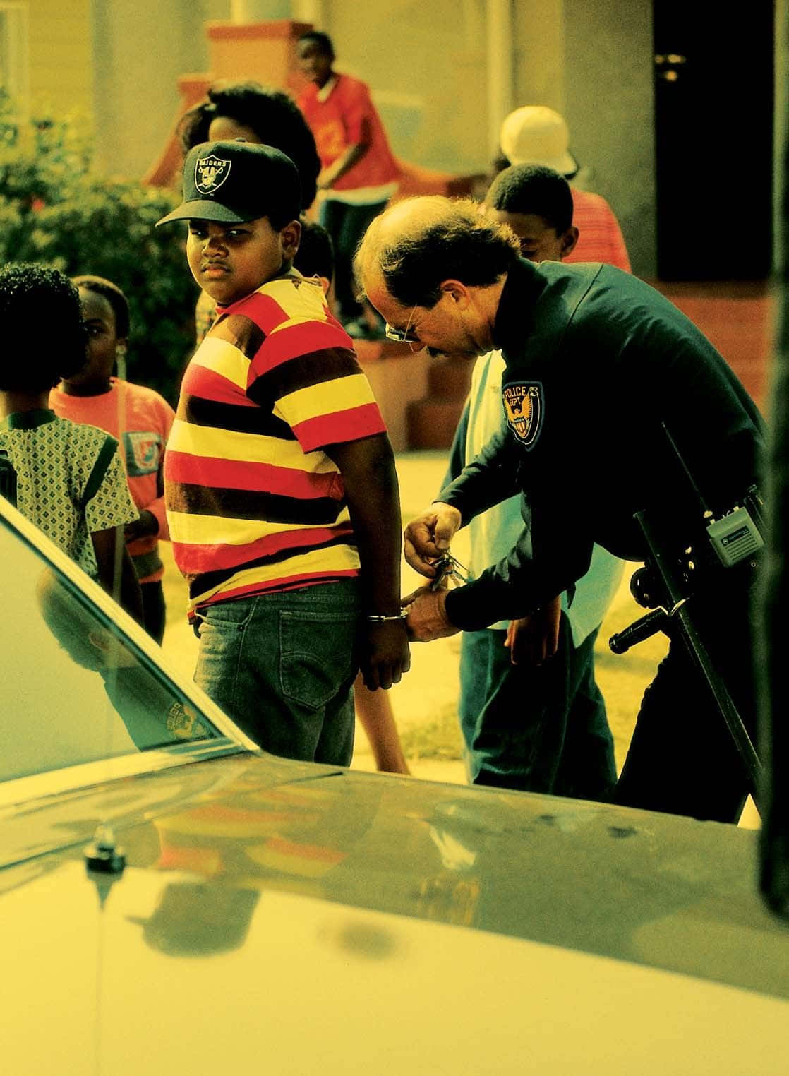 重溫《街区男孩》的傳奇遺產 壁紙 — 一名警察在陽光明媚的城市街道上，在汽車旁邊給一名穿著條紋襯衫的年輕人戴上手銬，旁觀者正在觀看。