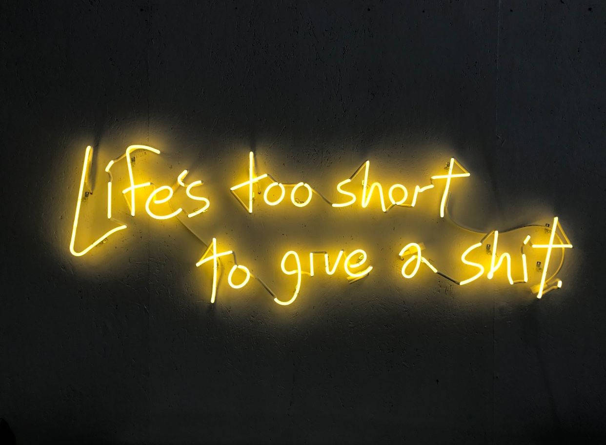 野性提醒霓虹黃 壁紙 — 一個發光的黃色霓虹燈牌，上面寫著 Life's too short to give a shit，安裝在深色紋理牆上。