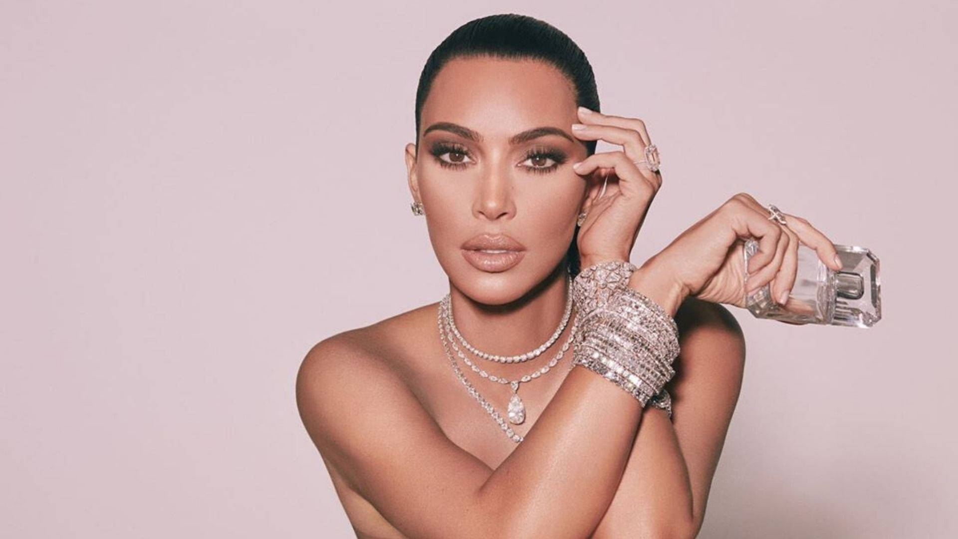 Kim Kardashian 手持香水瓶，佩戴閃亮珠寶，背景為粉紅色。