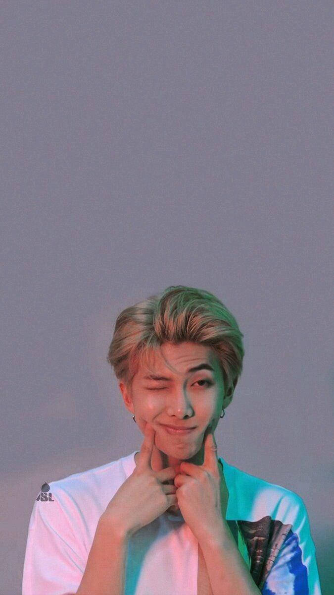 金南俊 Love Yourself 壁紙 — 一位金髮年輕男子在彩色霓虹燈光下，雙手捧著臉頰，俏皮地眨眼。
