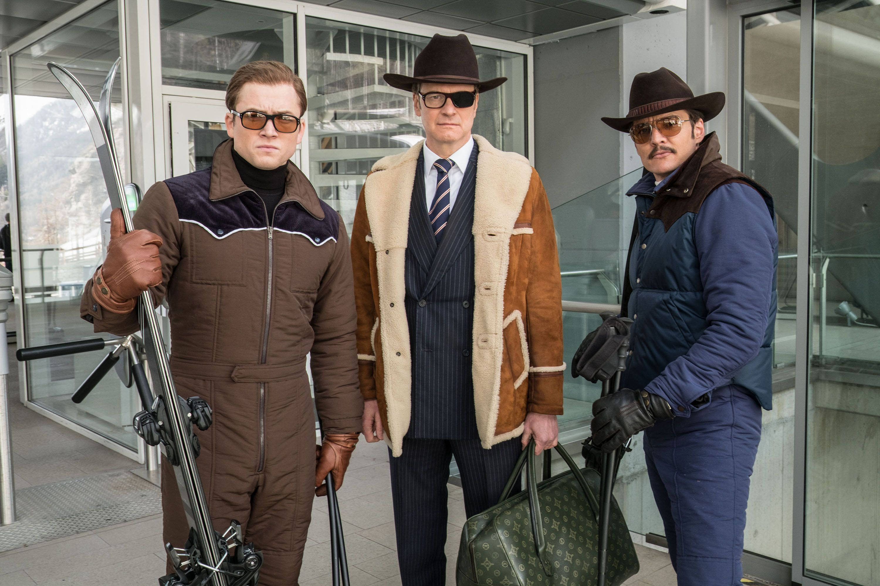 金士曼 Eggsy、加拉哈德與威士忌 壁紙 — 三位身穿時尚冬季裝備的男士自信地站在現代玻璃建築外，手持滑雪板和設計師手袋。