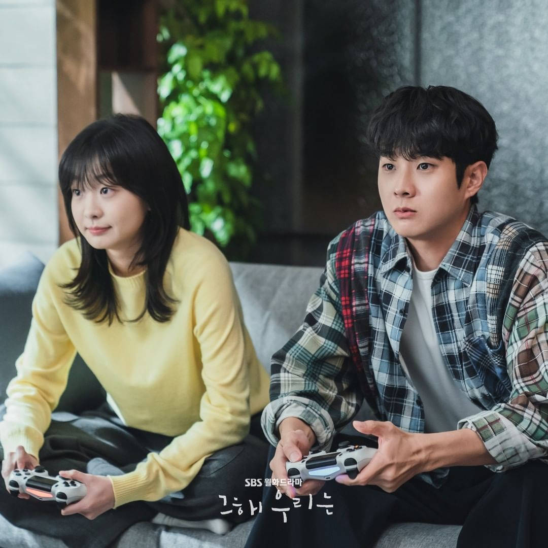 金多美與崔宇植玩PlayStation 壁紙 — 一對年輕情侶坐在沙發上，手持遊戲控制器，專注於屏幕，背景有柔和自然光線和綠色植物。