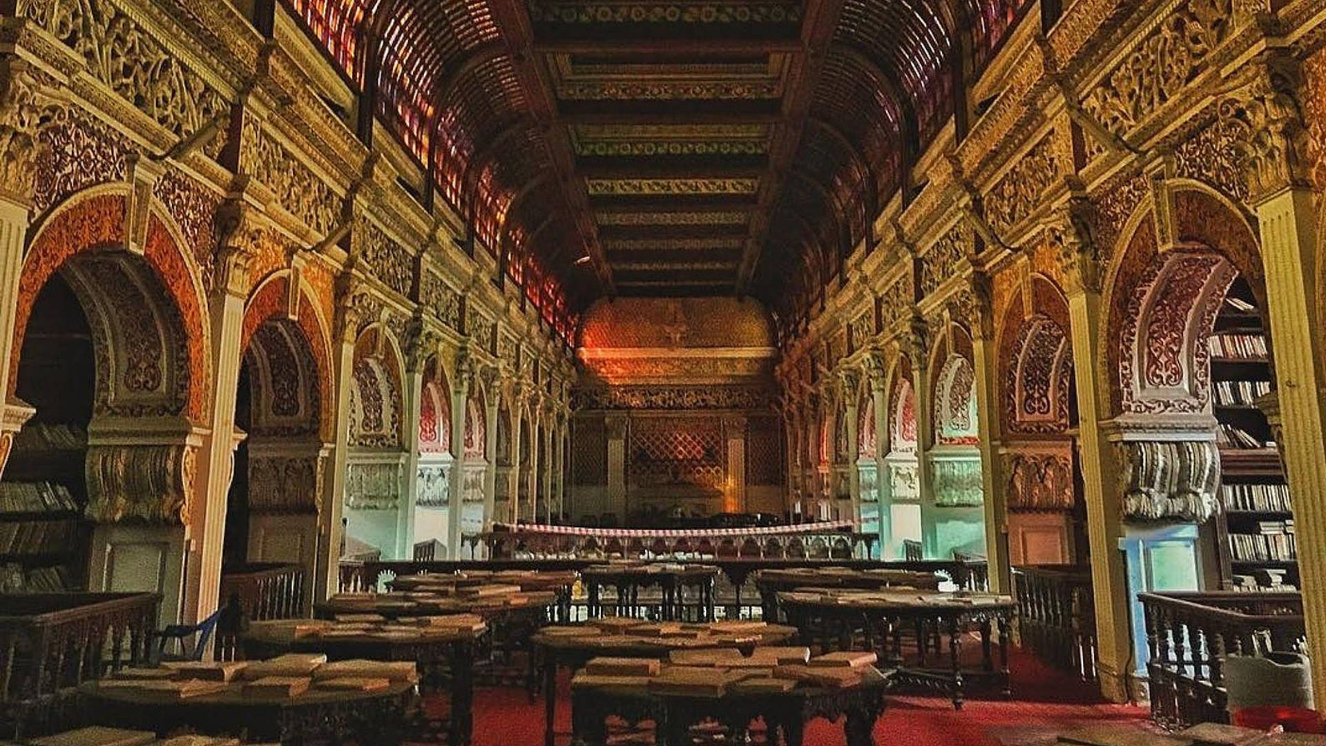 金奈康內馬拉圖書館 壁紙 — 宏偉華麗的圖書館，設有拱形書架、精緻雕刻和成排的木桌，在溫暖的金色燈光下。