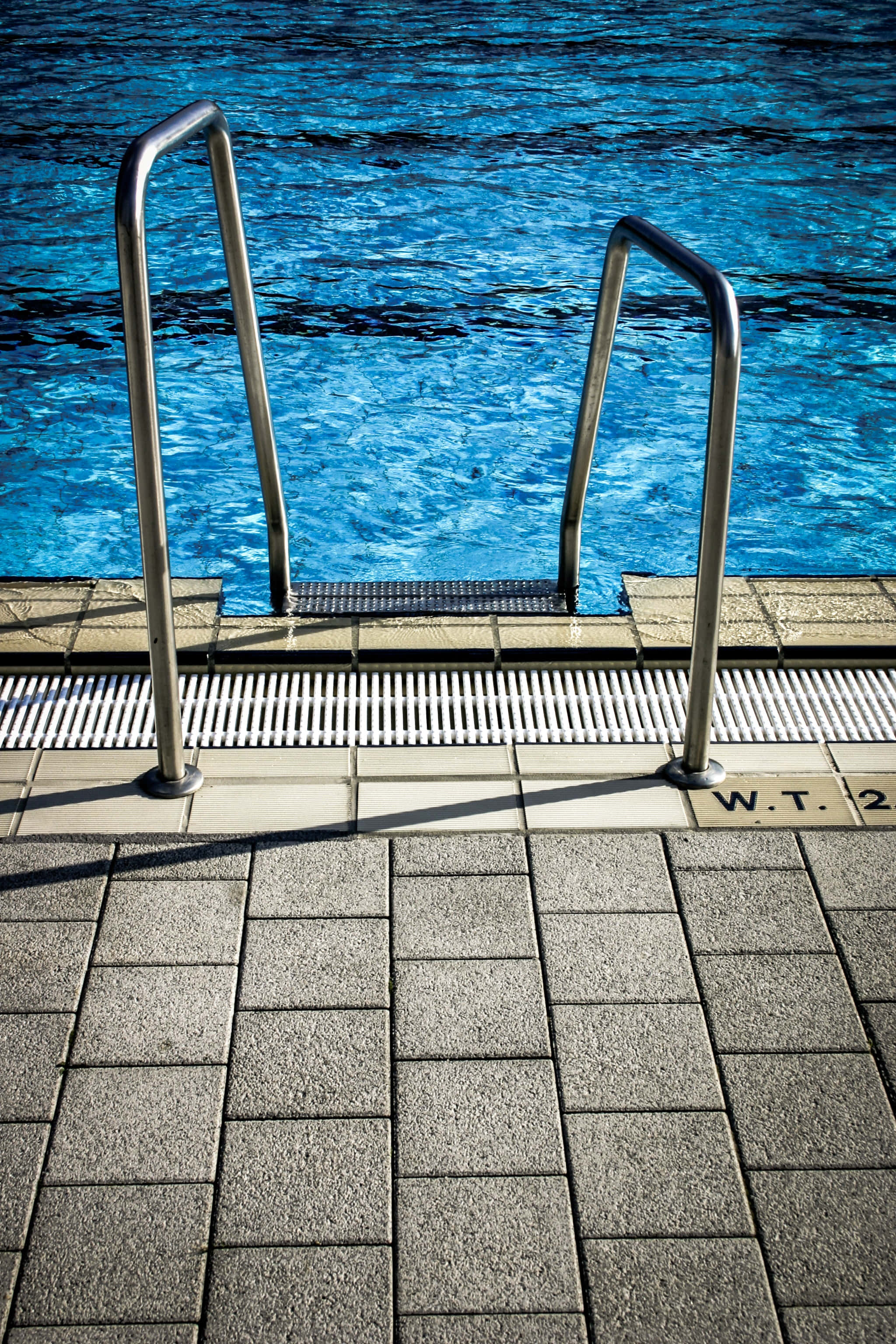 Swimming Pool 桌布 壁紙 — 金屬泳池梯子，帶有紋理踏板，立於陽光普照的瓷磚甲板上，旁邊是充滿活力的藍色水面。