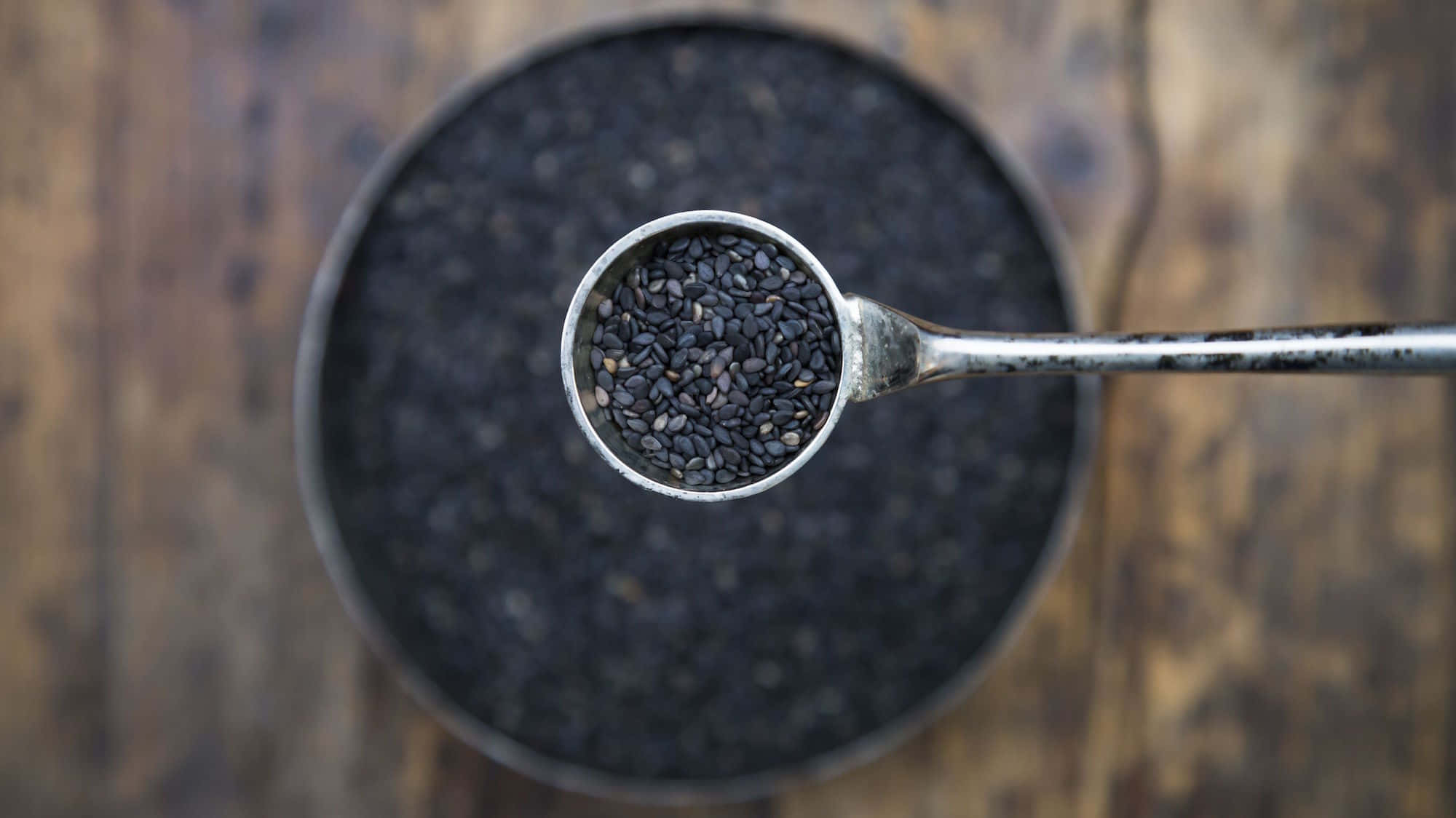 Black Sesame 桌布 壁紙 — 金屬湯匙將光澤的黑芝麻舀起，置於裝滿更多芝麻的質樸木碗上方。