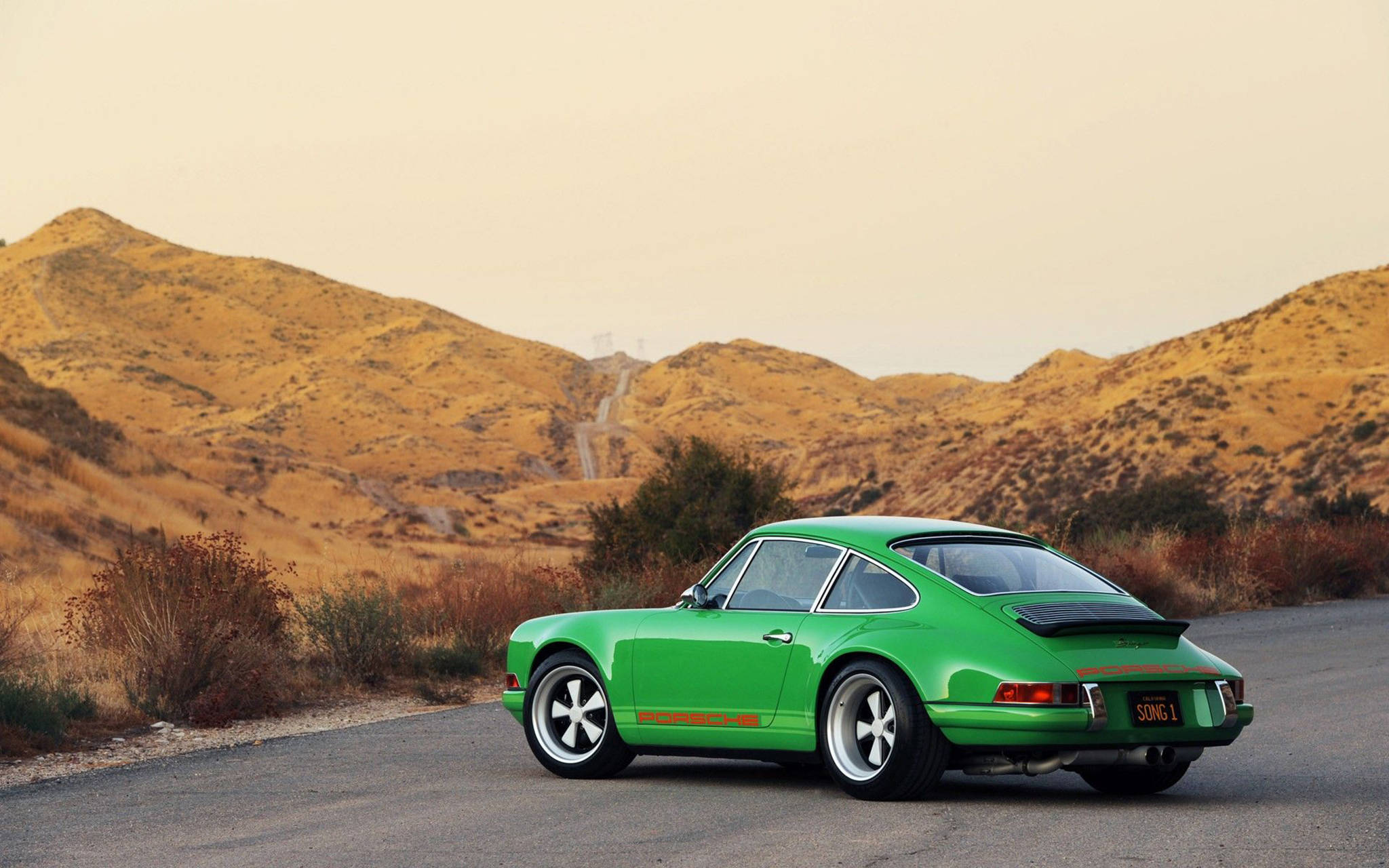 金屬綠色Singer Porsche 壁紙 — 一輛充滿活力的綠色經典保時捷911在朦朧的天空下，沿著兩旁金色山丘的沙漠公路行駛。