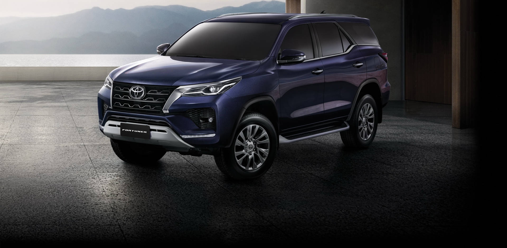 深藍色豐田 Fortuner SUV 停放在混凝土平台上，俯瞰山脈和海洋。