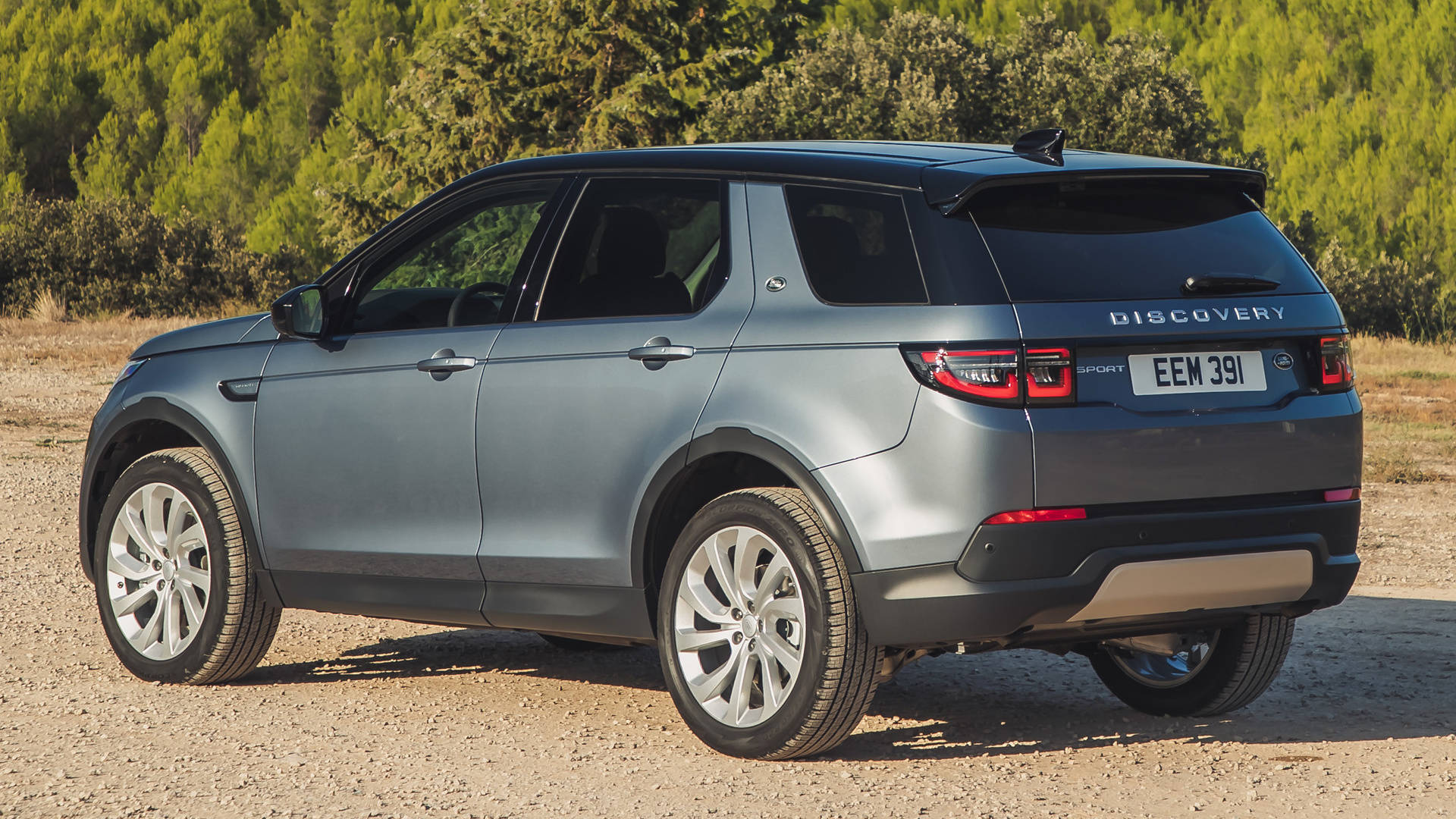 金屬藍色路虎 iPhone 主題壁紙 壁紙 — 一輛時尚的灰色 Land Rover Discovery Sport 停在碎石路上，背景是翠綠的樹木，在明亮的陽光下。