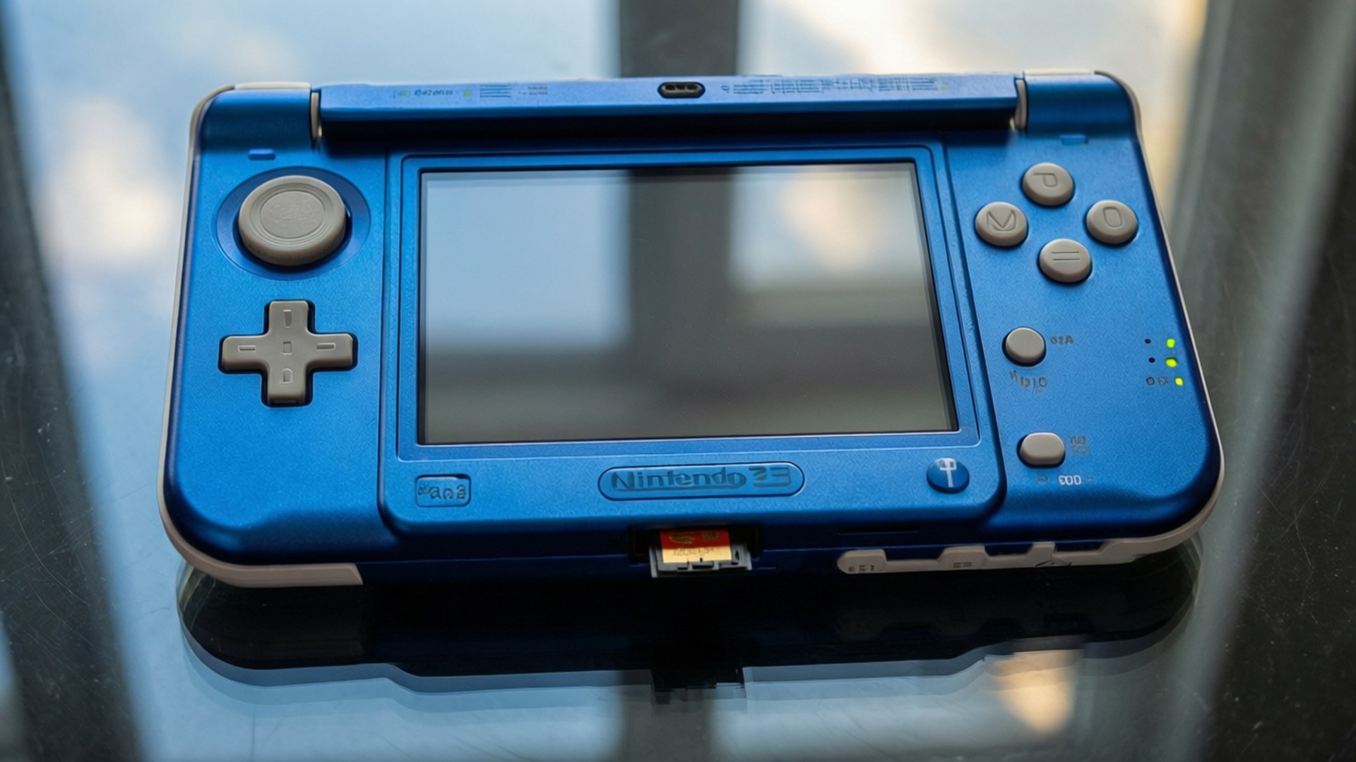 金屬藍頂視角 壁紙 — 一部藍色 Nintendo 3DS 手提遊戲機放置在反光玻璃表面上，屏幕關閉，綠色電源指示燈亮起。