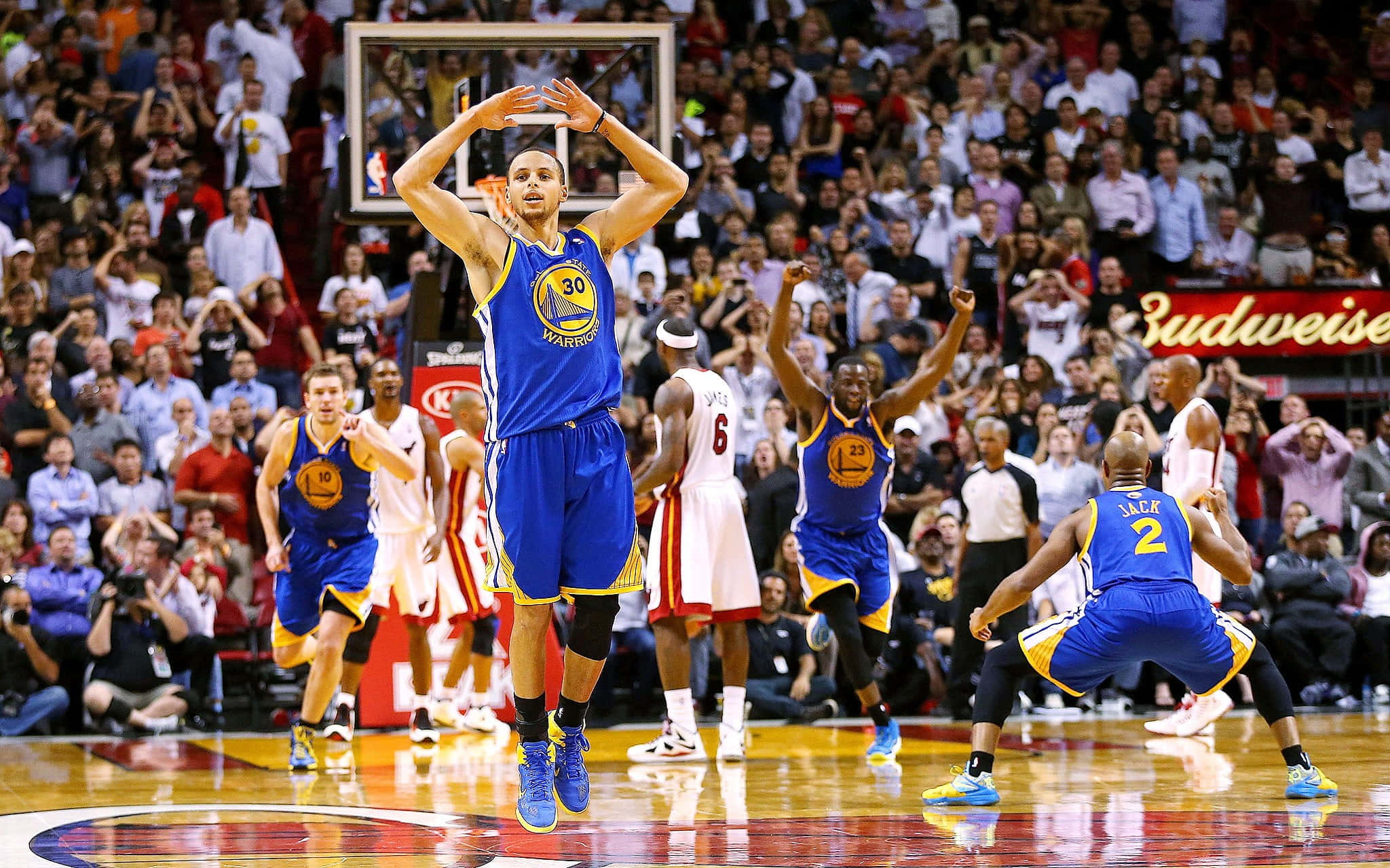 steph curry 桌布 壁紙 — 金州勇士隊球員身穿藍色球衣，在空中跳起投籃，隊友和觀眾在擠滿的場館中歡呼。