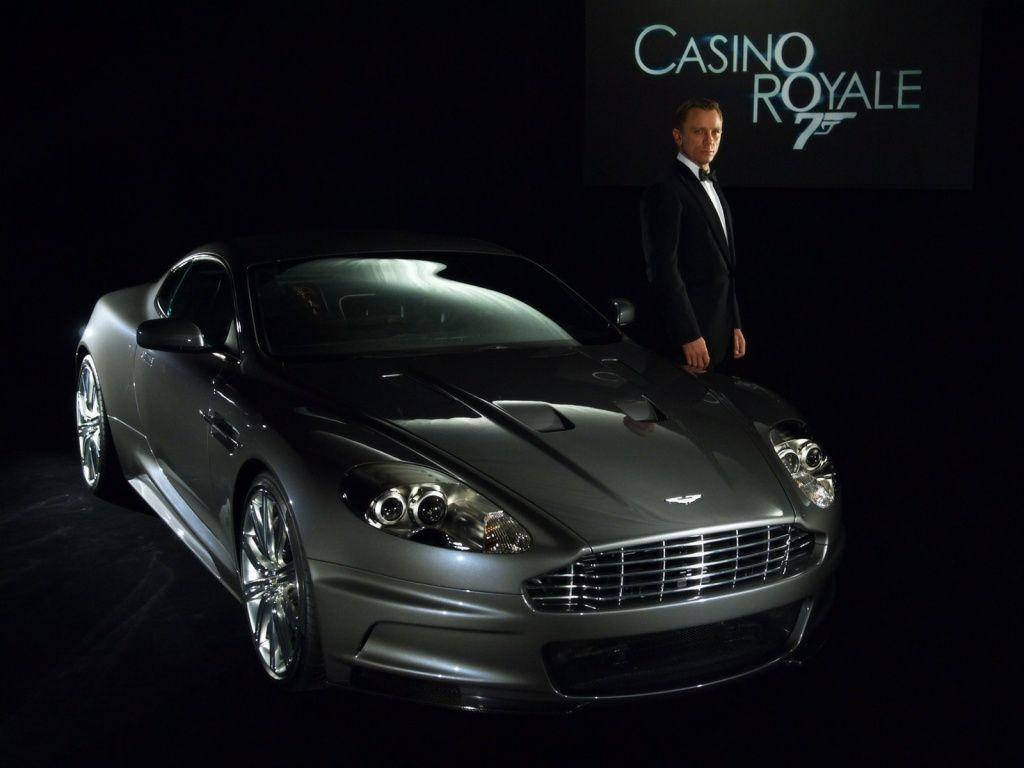 金手指黑色豪華車 壁紙 — 一架流線型的銀色Aston Martin DBS在攝影棚燈光下閃閃發光，丹尼爾·基克身穿禮服站在旁邊，背景為黑色，帶有Casino Royale 75標誌。