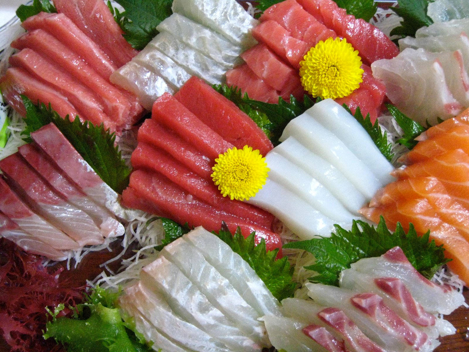 金槍魚、三文魚、鰹魚刺身 壁紙 — 一個色彩繽紛的雜錦刺身拼盤，有粉紅色的吞拿魚、白色的魚肉和橙色的三文魚片，並以黃色花朵和綠色葉子作裝飾。
