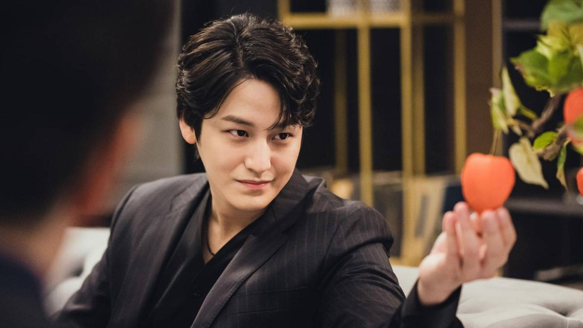 金汎凝視橙果 壁紙 — 一名身穿深色西裝的男子手持紅色心形物體，同時看著另一個人，場景在柔和燈光的室內環境中。