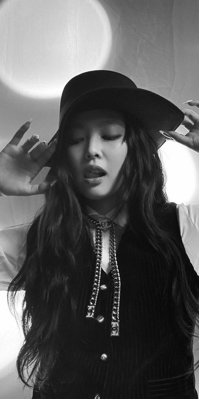 金珍妮潮流酷感造型 壁紙 — 一位戴著寬邊帽的女性，雙手靠近臉龐，以柔和光線下的紋理牆壁為背景，呈現單色調。