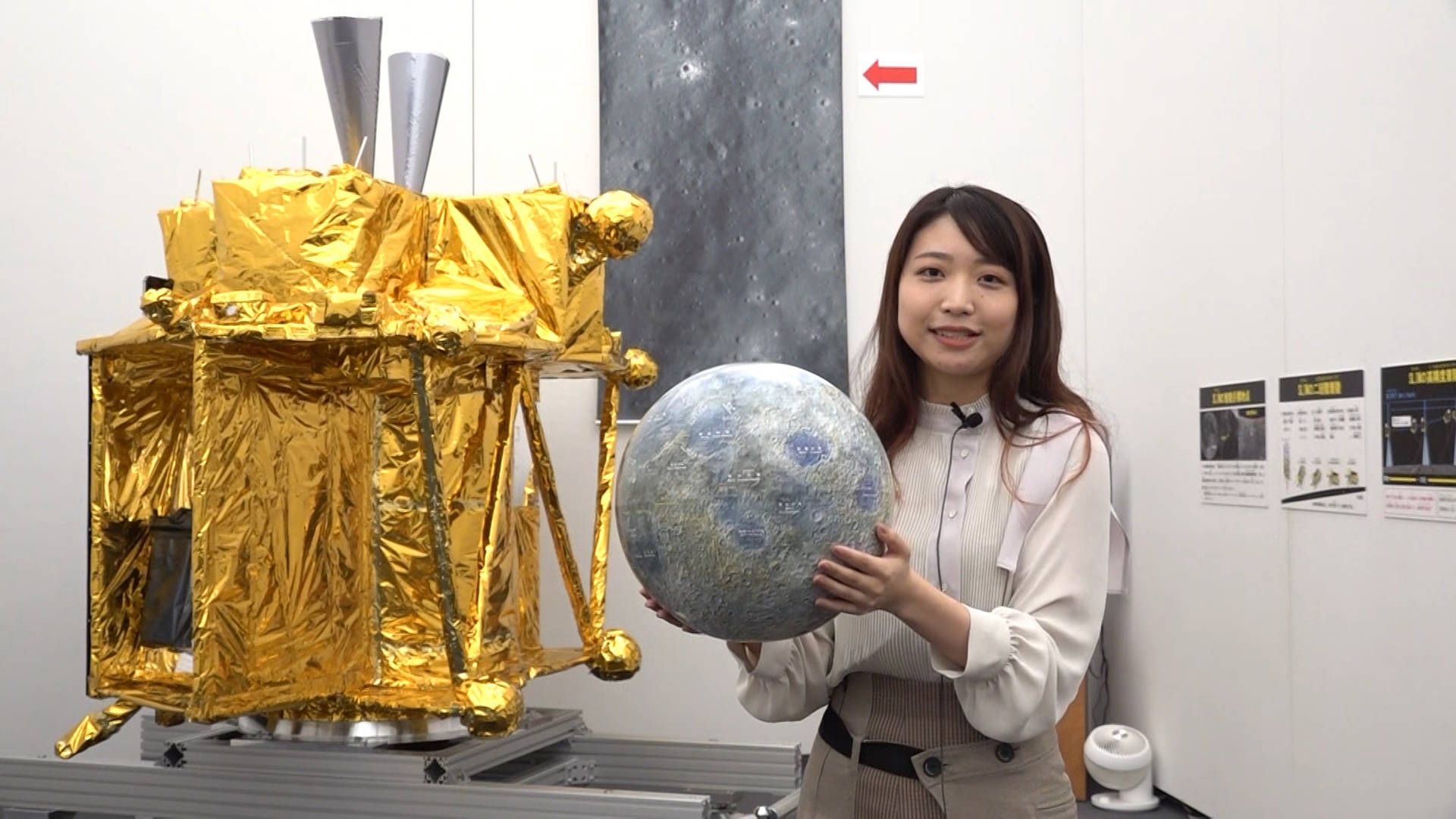 金箔學校專案 壁紙 — 一位女士在潔白的展覽空間中微笑，手持月球模型，旁邊是鍍金箔的太空船複製品。