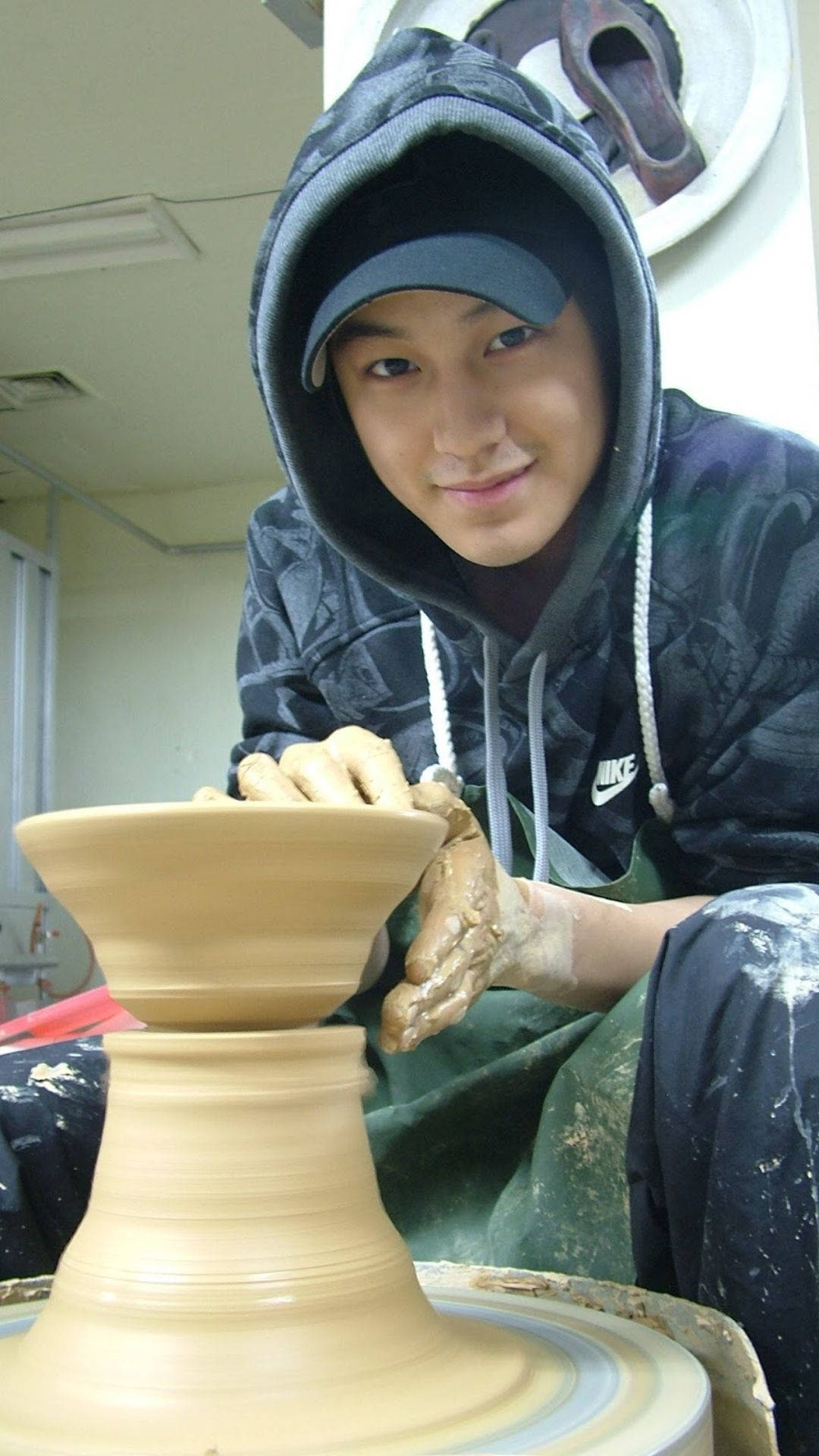 金範陶藝師 壁紙 — 一位穿著連帽衫的年輕男子在工作室中微笑著在陶輪上塑造濕黏土。
