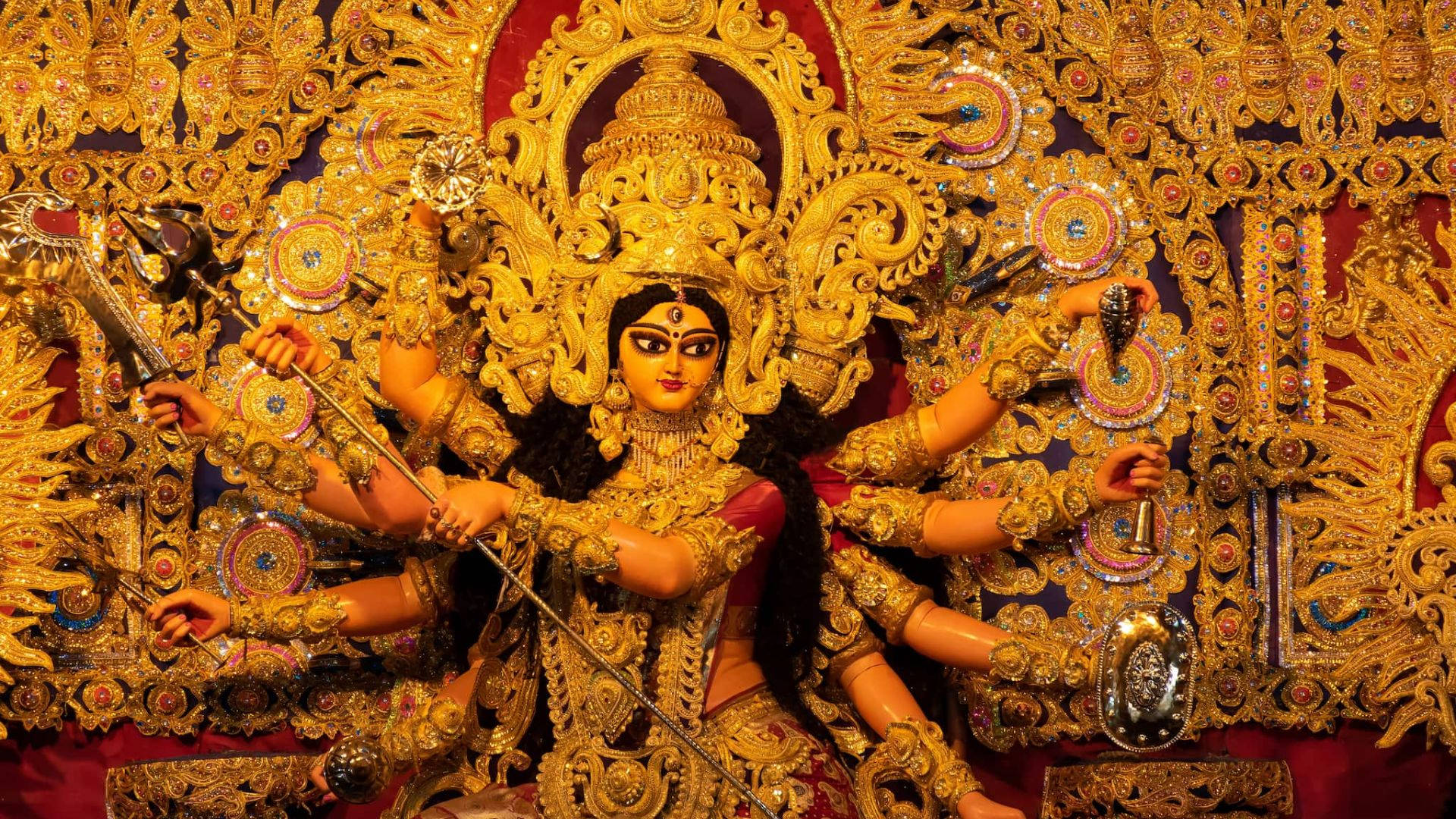 金色 Durga Mata 高清 壁紙 — 在珠寶背景前，一尊閃耀的金色杜爾迦神像，多臂手持武器。