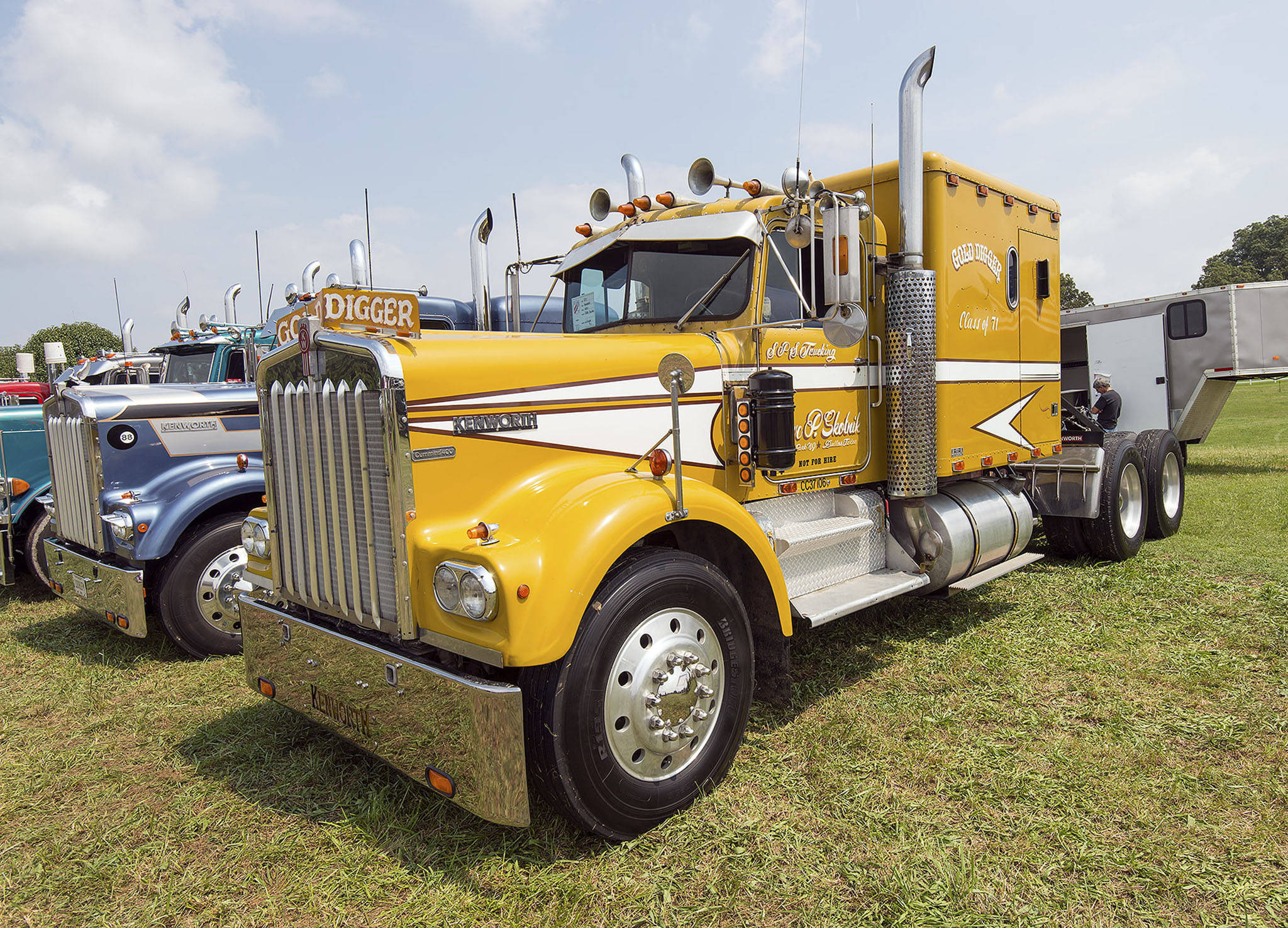 金色Kenworth W900 壁紙 — 一輛鮮黃色經典半掛卡車，帶有鍍鉻細節和定制貼花，停在卡車展的草地上，天空部分多雲。