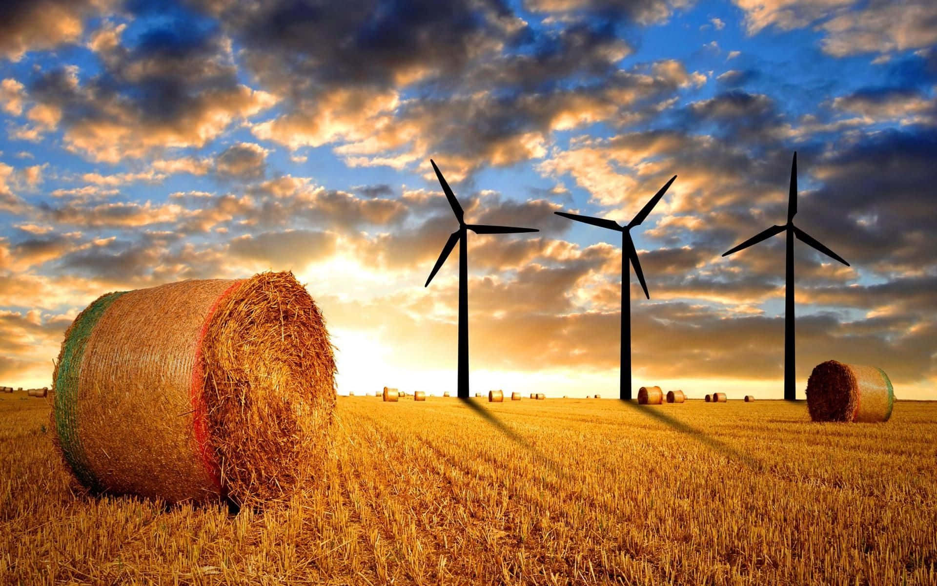 Energy 桌布 壁紙 — 金色乾草捆點綴在陽光照耀的田野上，風力發電機在戲劇性的日落天空下投下長長的影子。