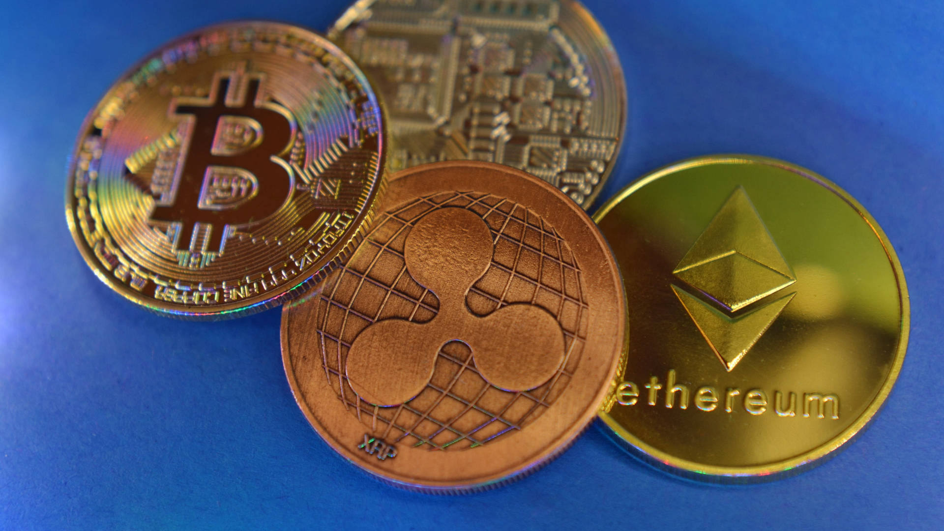 金色以太幣硬幣 壁紙 — 四個實體加密貨幣硬幣 Bitcoin Ethereum XRP 和 Litecoin 在明亮燈光下放置在充滿活力的藍色表面上。
