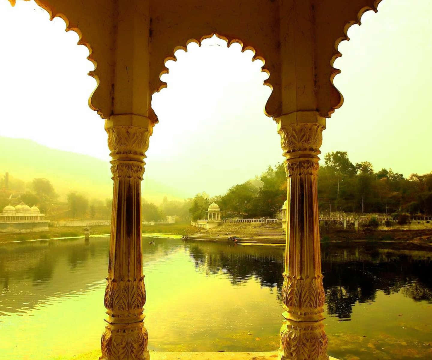 Rajasthan 桌布 壁紙 — 金色光線穿過華麗的柱子，框住一條寧靜的河流，遠處的寺廟倒影在迷霧的早晨薄霧中。