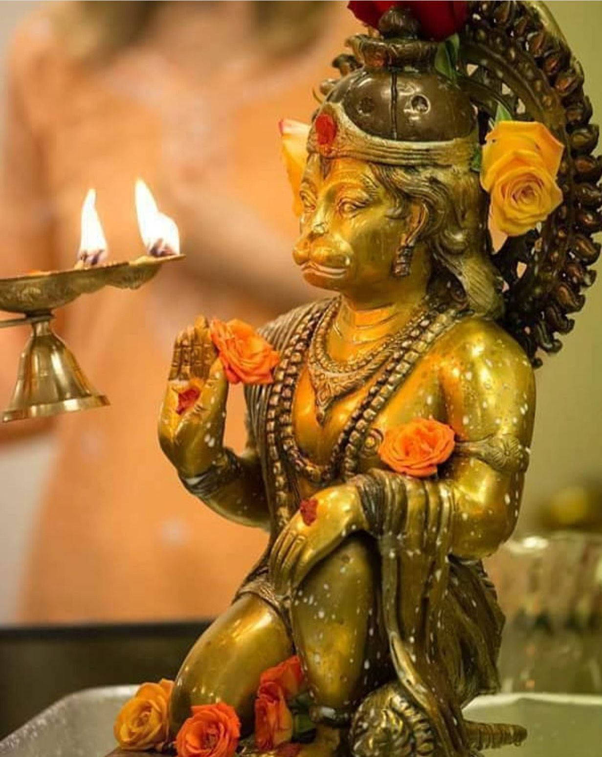 Indian 桌布 壁紙 — 金色哈努曼雕像飾以橙色玫瑰，並在寧靜的寺廟環境中手持點亮的油燈。