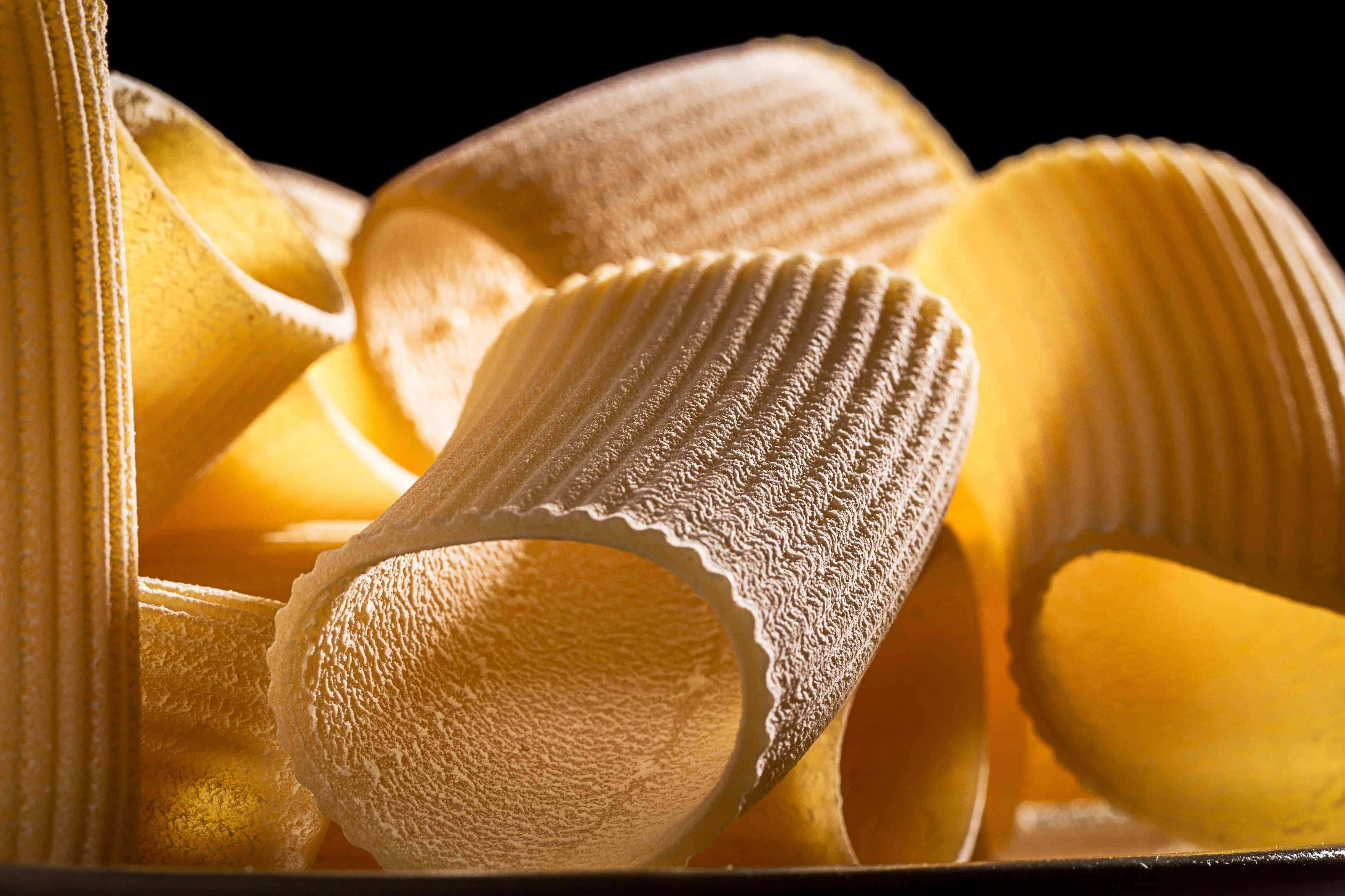 Pasta 桌布 壁紙 — 金色坑紋意粉管在溫暖燈光下閃閃發光，襯托著純黑背景。